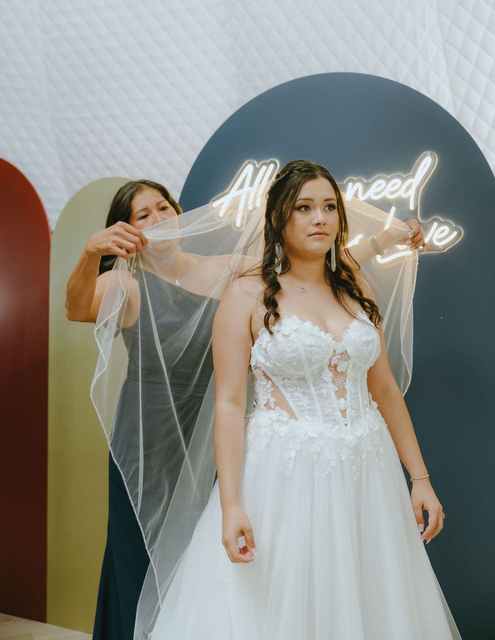 Bride in white lace ballgown and veil being adjusted by bridesmaid in front of neon sign.
