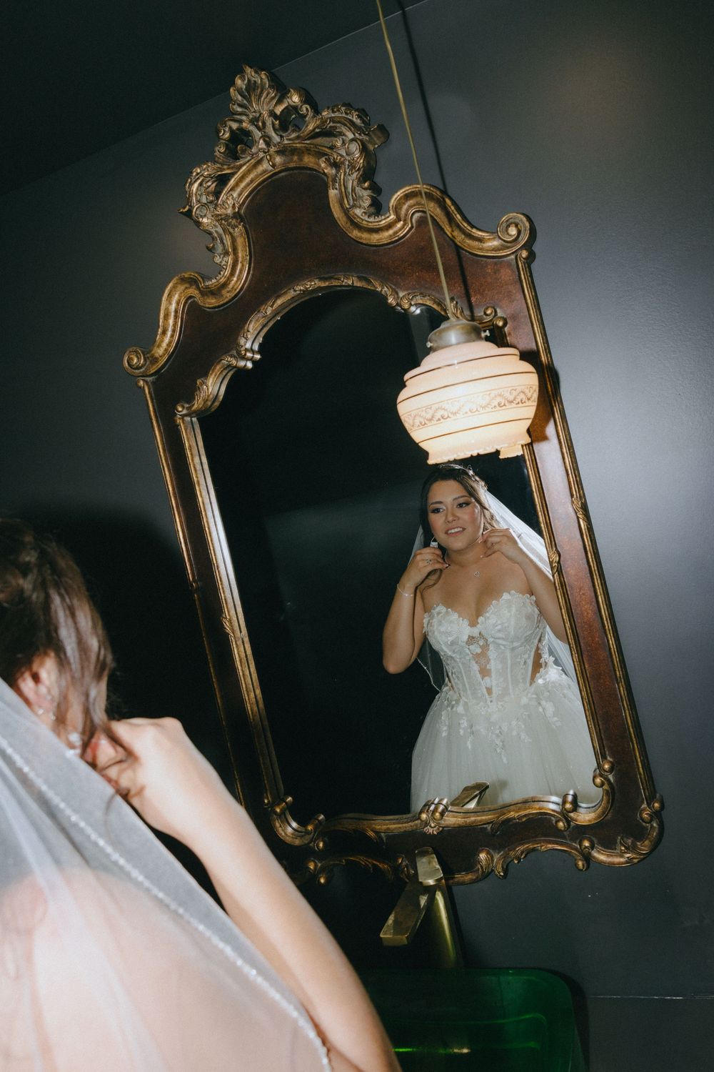 Bride in lace corset wedding gown admires her reflection in an ornate gold vintage mirror with a glowing pendant lamp.