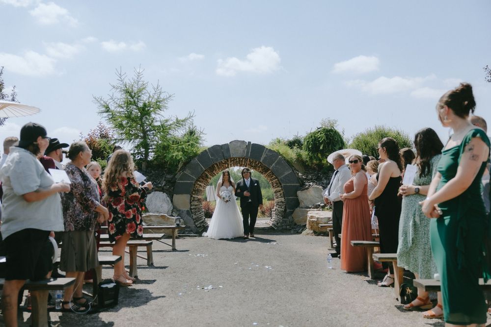 Outdoor wedding ceremony with guests standing along the aisle as bride and groom walk under a stone arch on a sunny day.