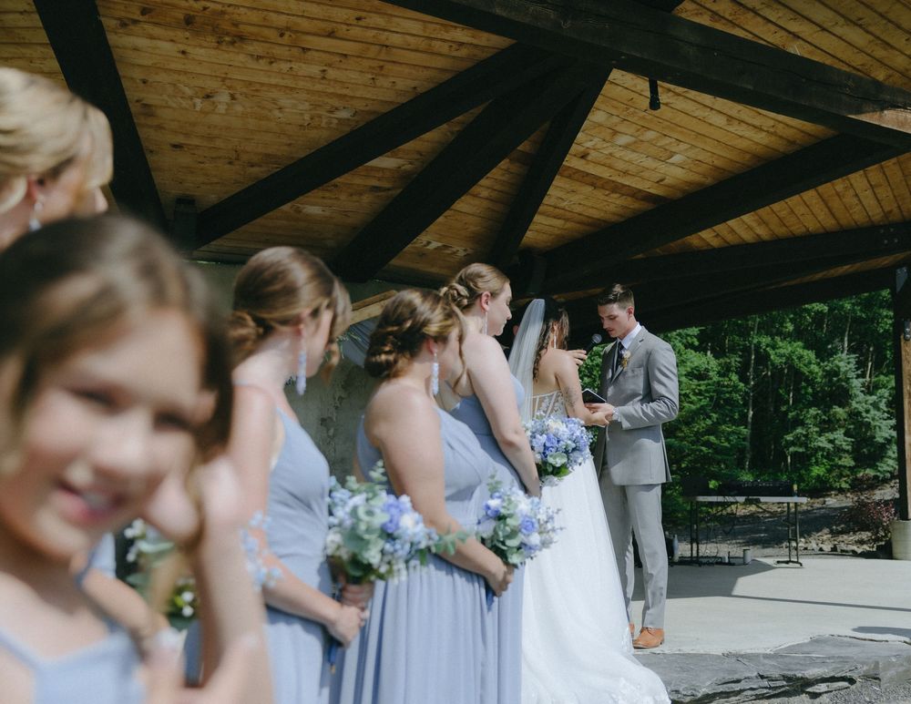 Bridesmaids in light blue dresses holding floral bouquets stand beside a bride and groom exchanging vows under a wooden pavilion.