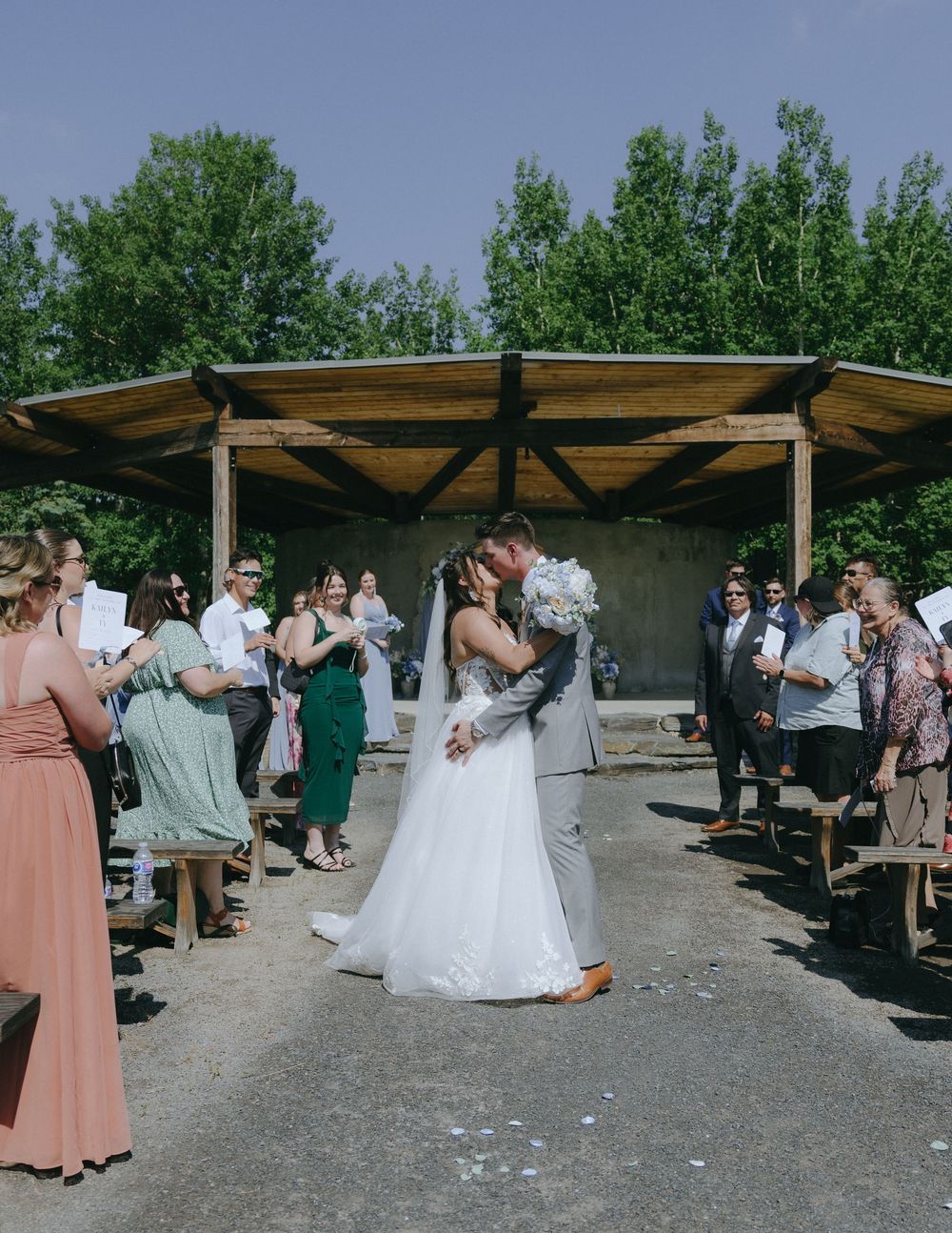 Bride and groom share first kiss as newlyweds walking down outdoor wedding aisle surrounded by guests.