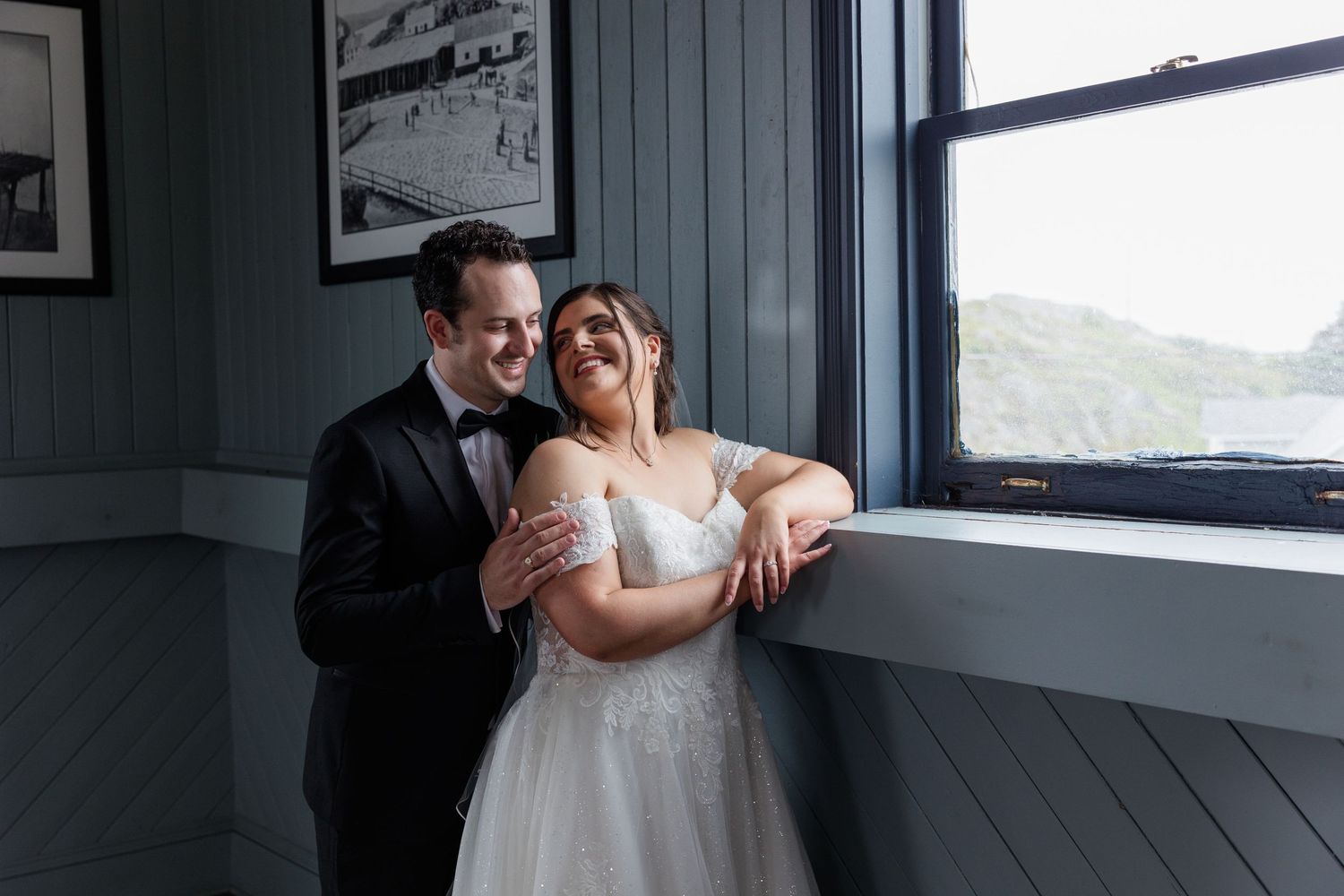 A bride and groom share a joyful moment by a window, she in a white gown and he in a black tuxedo.