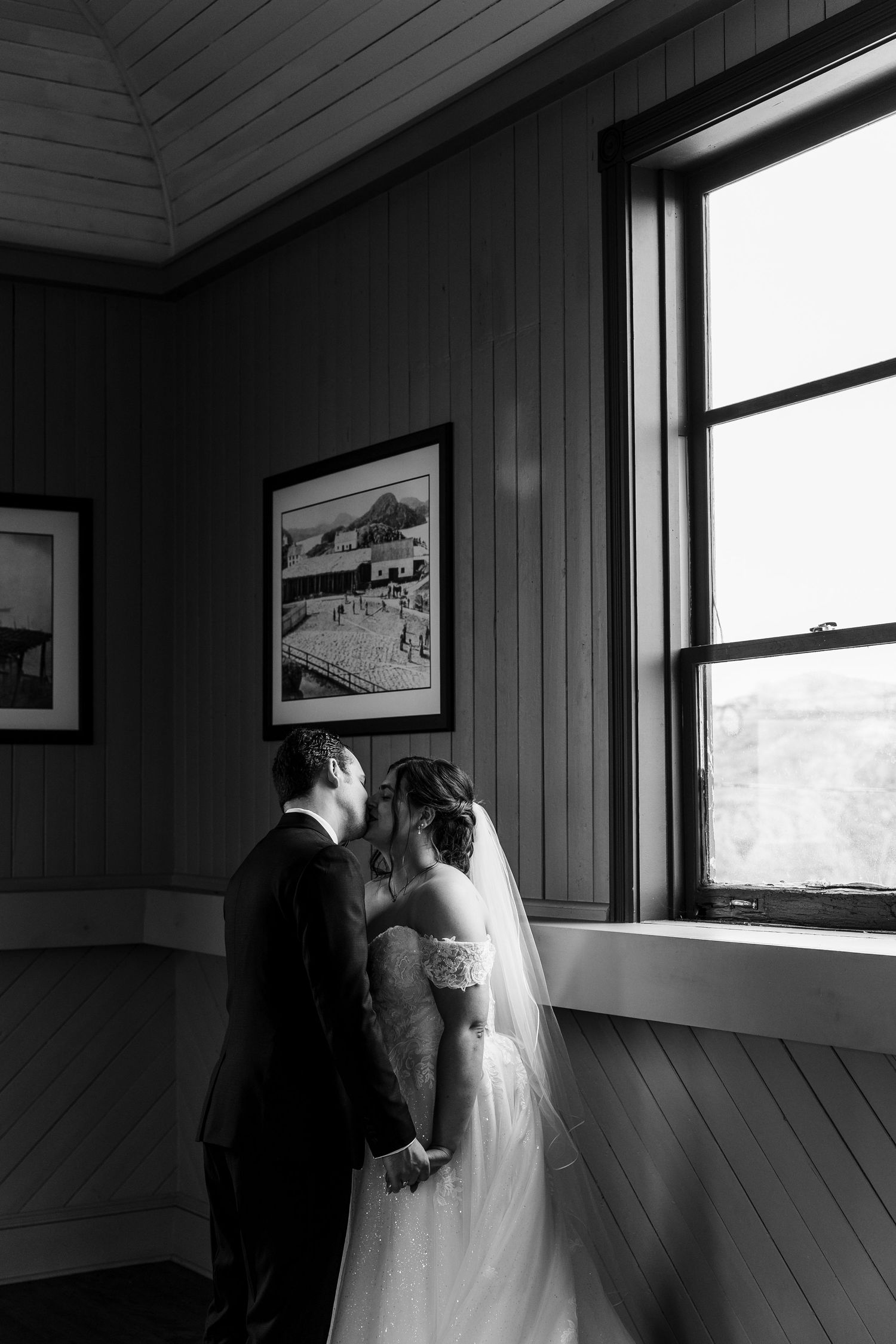 Black and white photo of a bride and groom sharing a kiss by a window in a rustic wood-paneled room on their wedding day.
