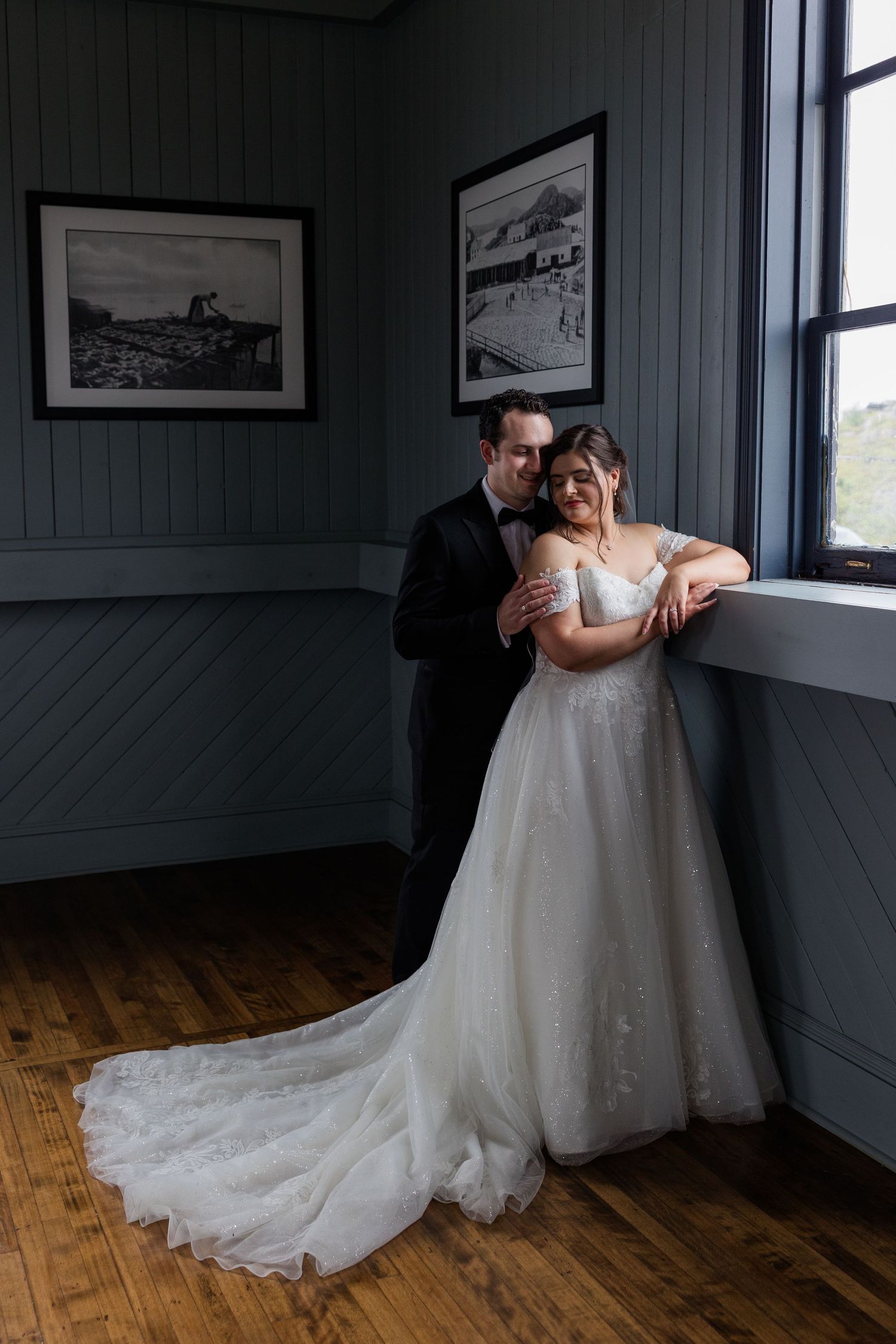 Bride and groom share a romantic embrace by a window, she in a flowing white gown, he in a classic black tuxedo.