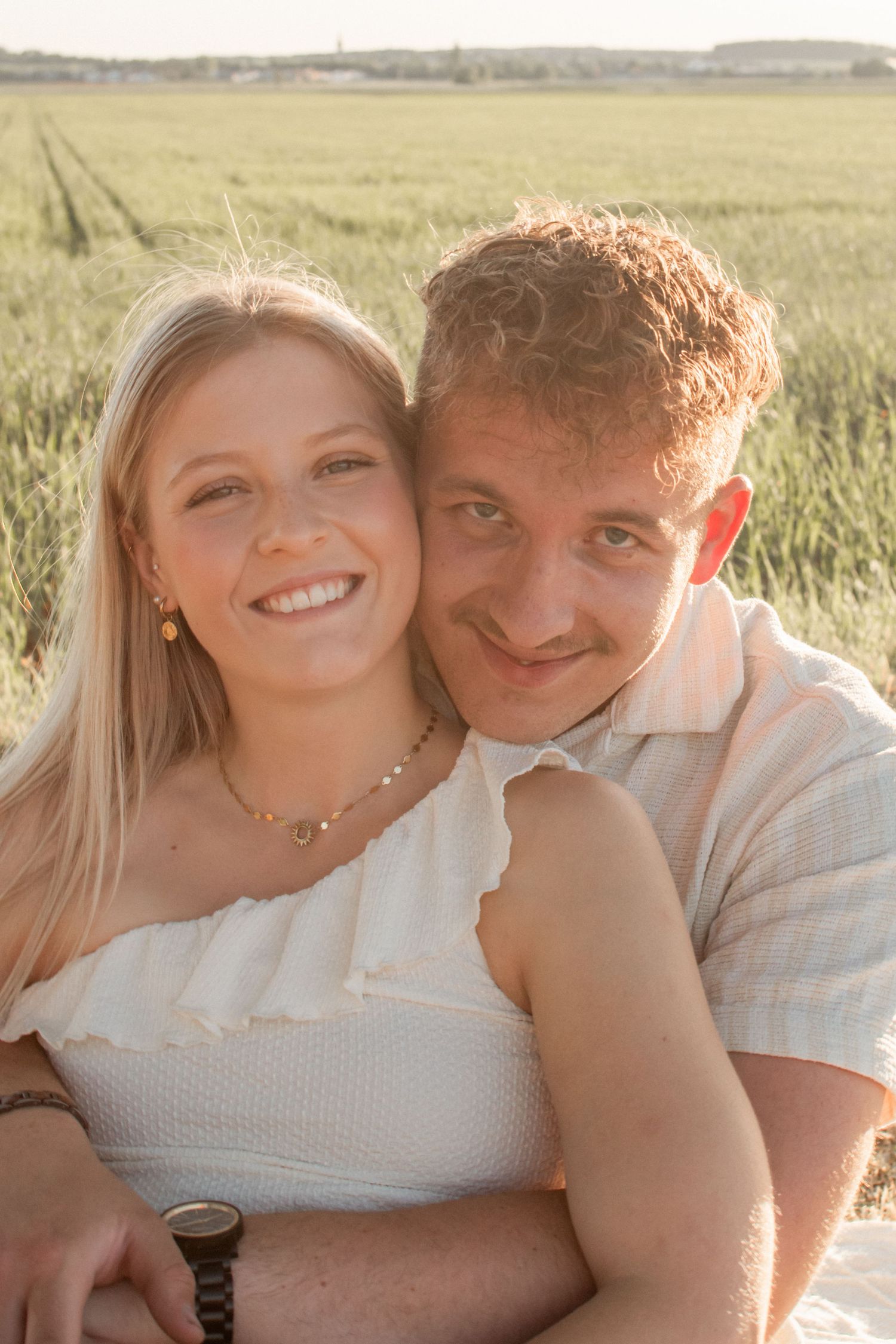 A smiling couple poses together in a sunny green field, the woman in white and the man in a light shirt.