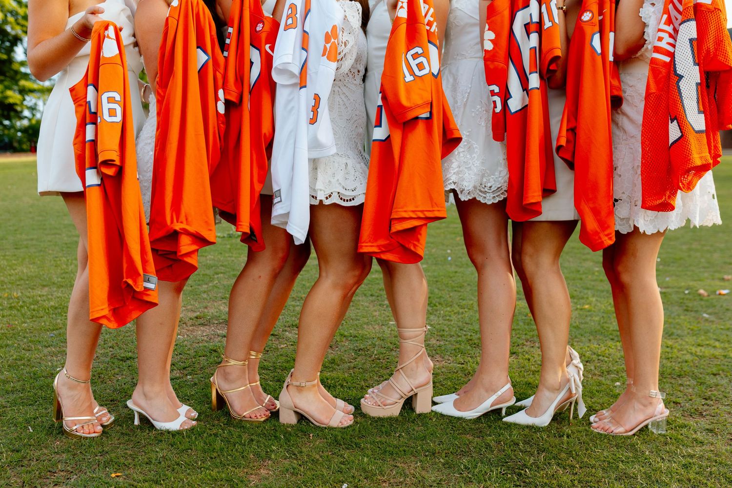 Bridesmaids holding orange Clemson football jerseys while wearing white dresses and nude heels on green grass.