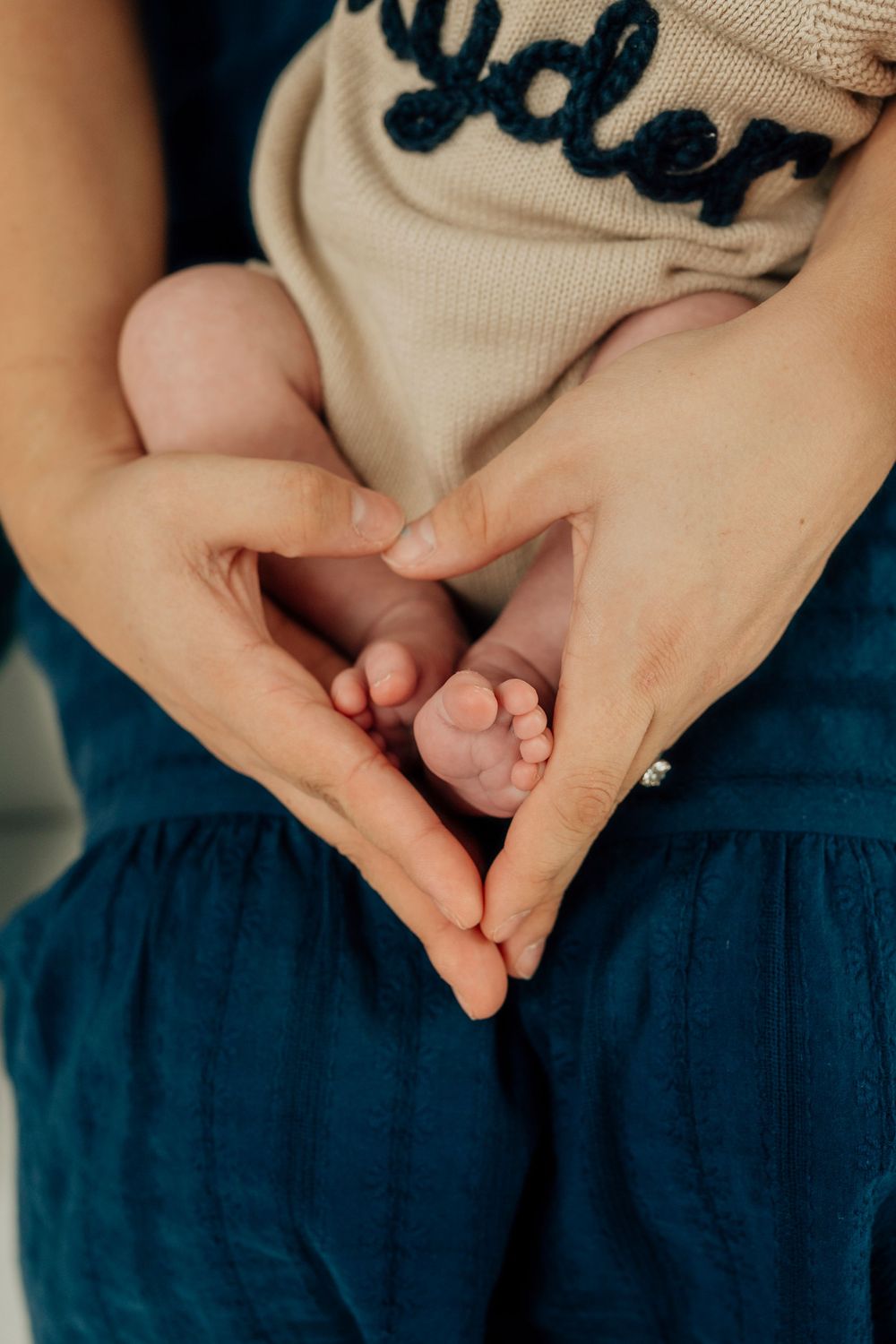 Mother cradling newborn baby feet in heart-shaped hands, wearing teal skirt and baby in beige outfit.