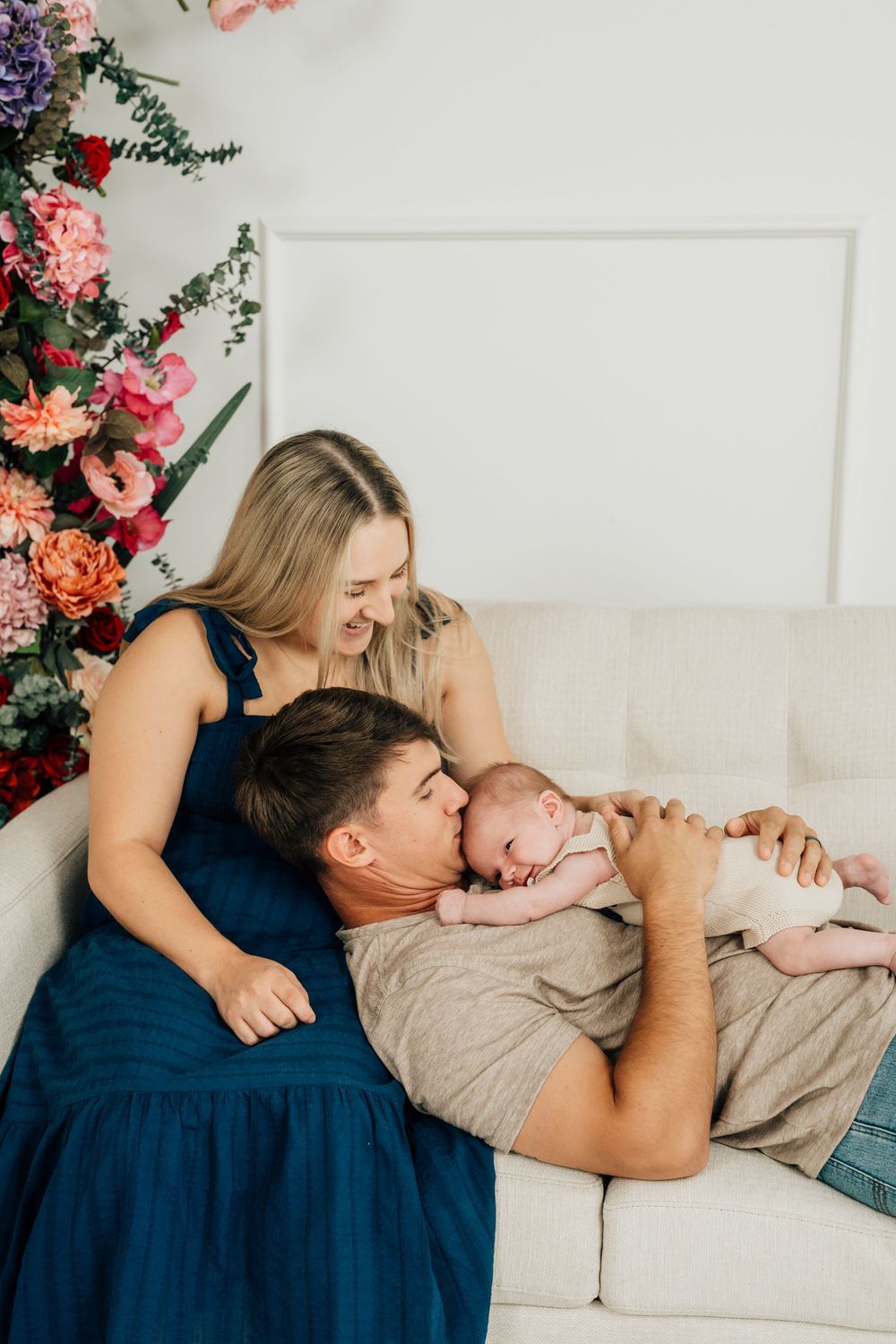 New parents tenderly kiss their newborn baby on a white sofa surrounded by colorful floral arrangements.