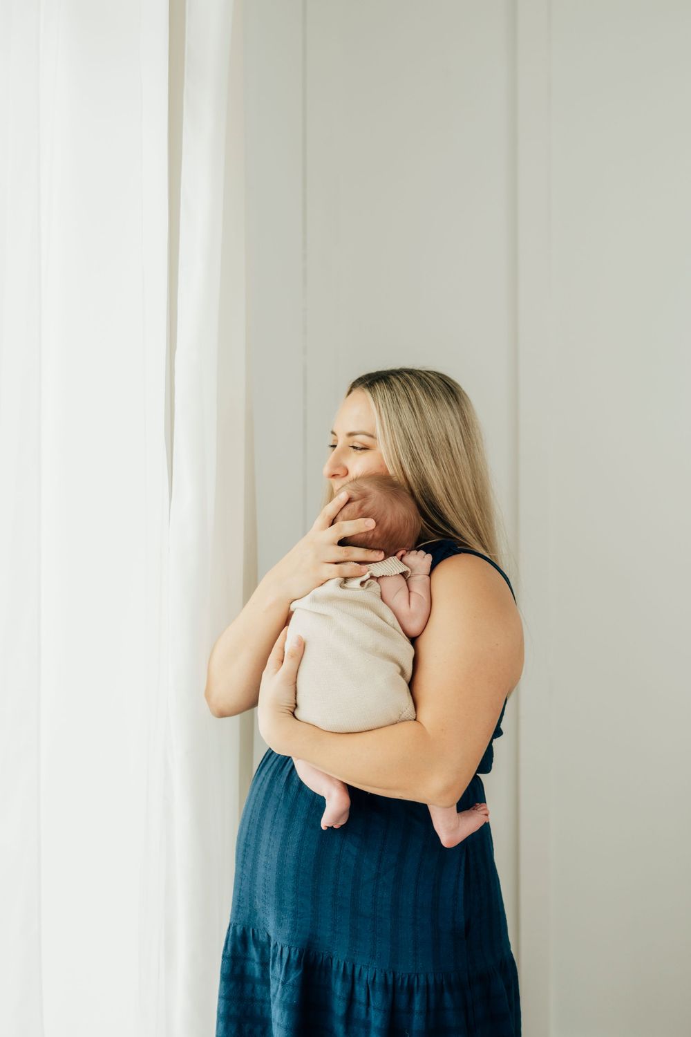 A mother in a blue dress tenderly kisses her newborn baby near a bright window.