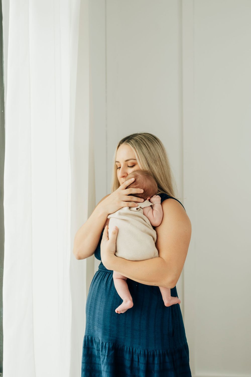 A blonde mother in a blue dress tenderly kisses her newborn baby wrapped in a cream swaddle near a bright window.