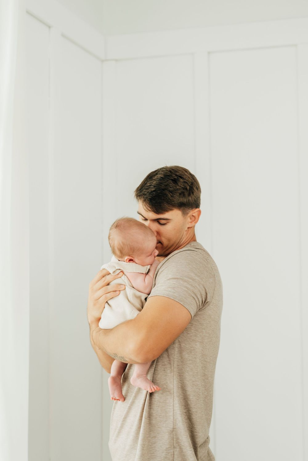 A young father in a gray t-shirt tenderly kisses his newborn baby while holding them against a white background.