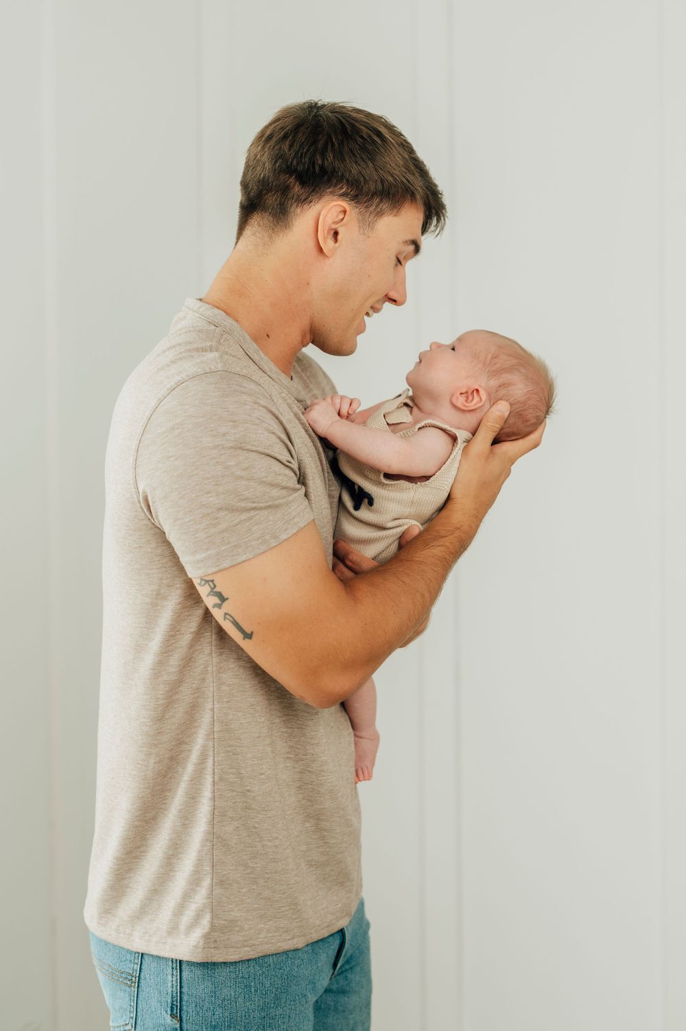 Young father in beige t-shirt lovingly holds his newborn baby, gazing at each other in a bright studio setting.