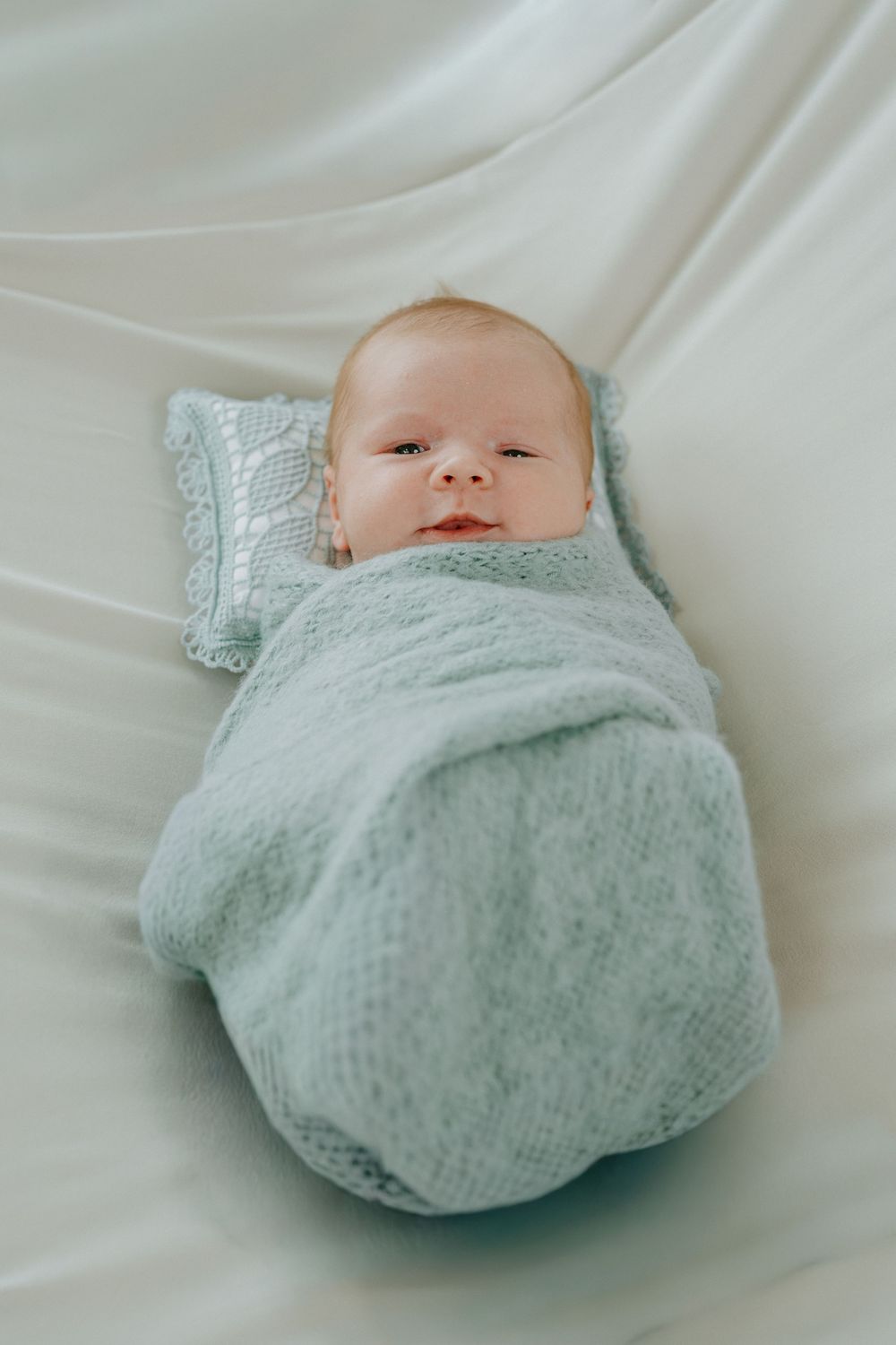 Newborn baby swaddled in a soft light blue knit blanket lying on white fabric.
