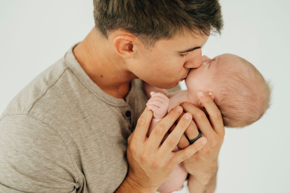 A loving father gently kisses his newborn baby, cradling the infant tenderly in both hands against a white background.