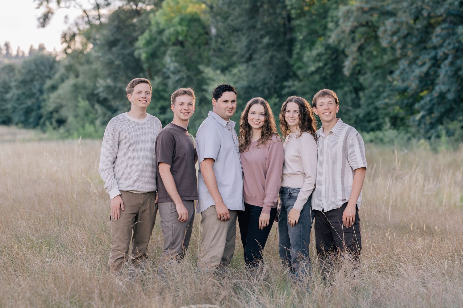 Six adults smiling together in a golden field with lush green trees in the background during a family portrait session.
