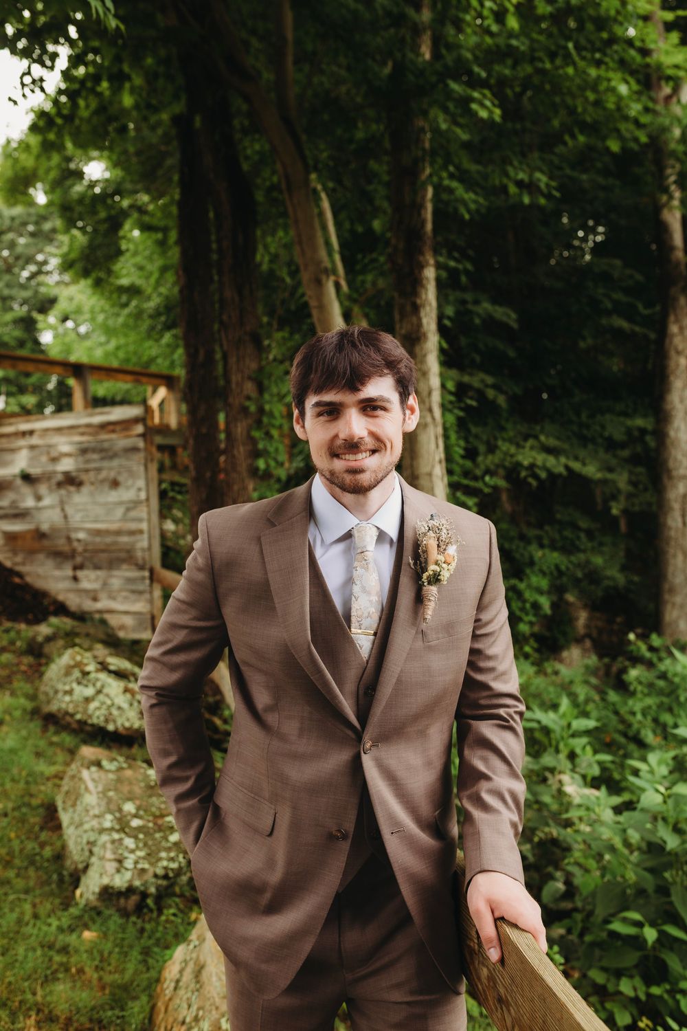 A groom in a brown three-piece suit with boutonniere smiles outdoors among lush green trees and rustic wooden structures.