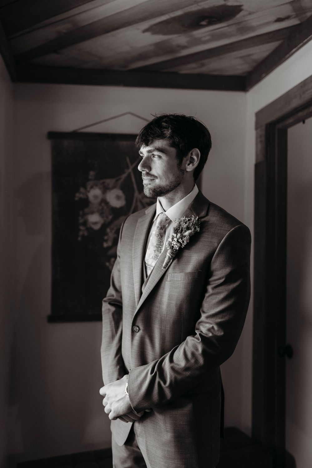 Groom in gray suit with boutonniere stands pensively in black and white wedding portrait.