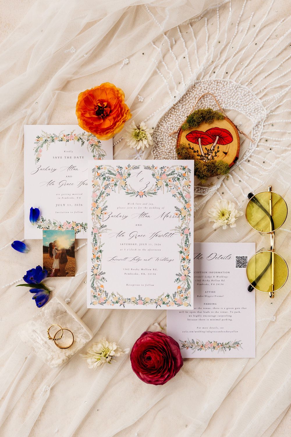 Elegant wedding invitation suite with floral details, gold rings, sunglasses, and colorful flowers on a white fabric backdrop.