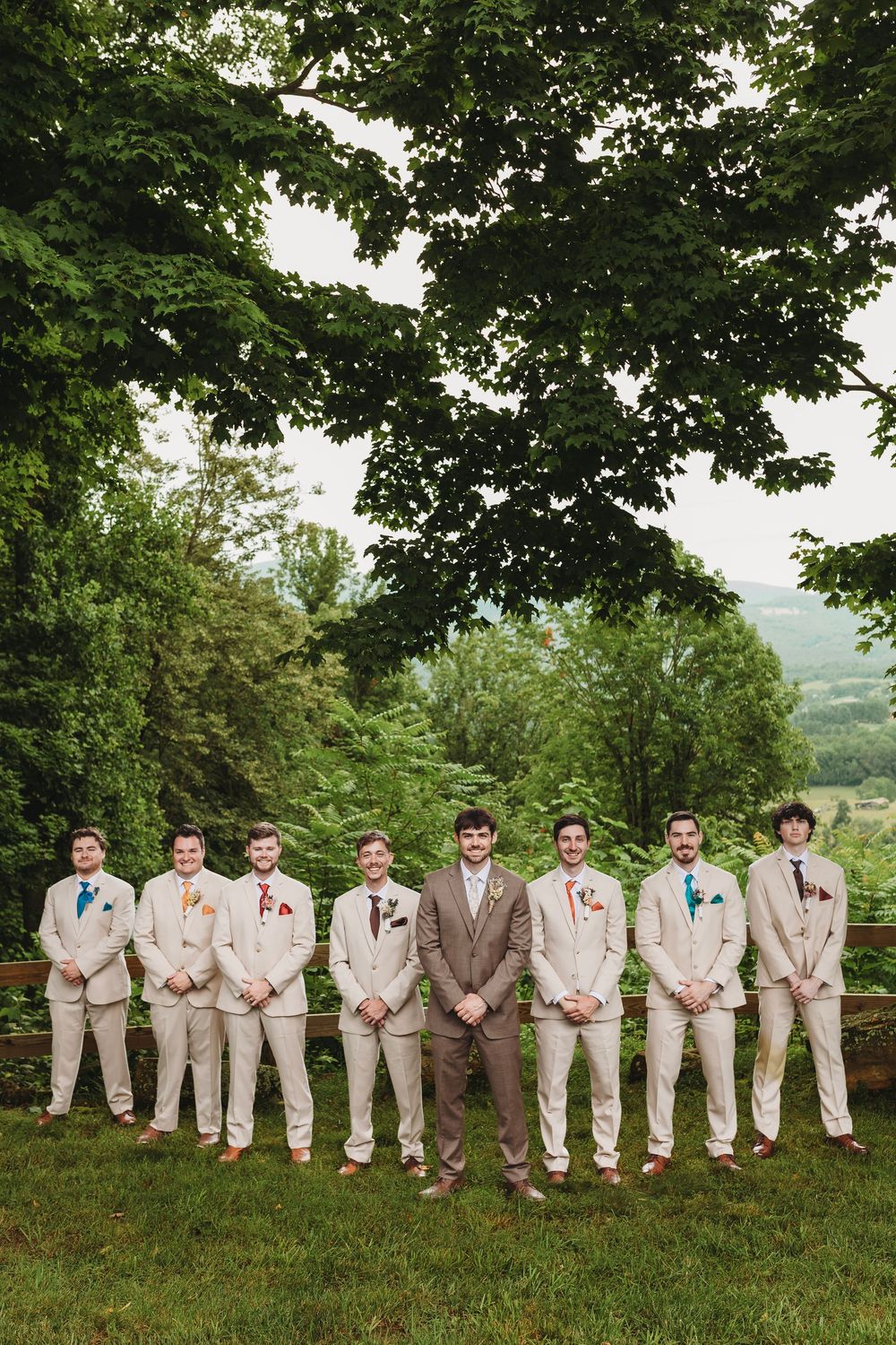 Groom and groomsmen in tan suits with colorful boutonnieres posing outdoors on green lawn with lush trees.