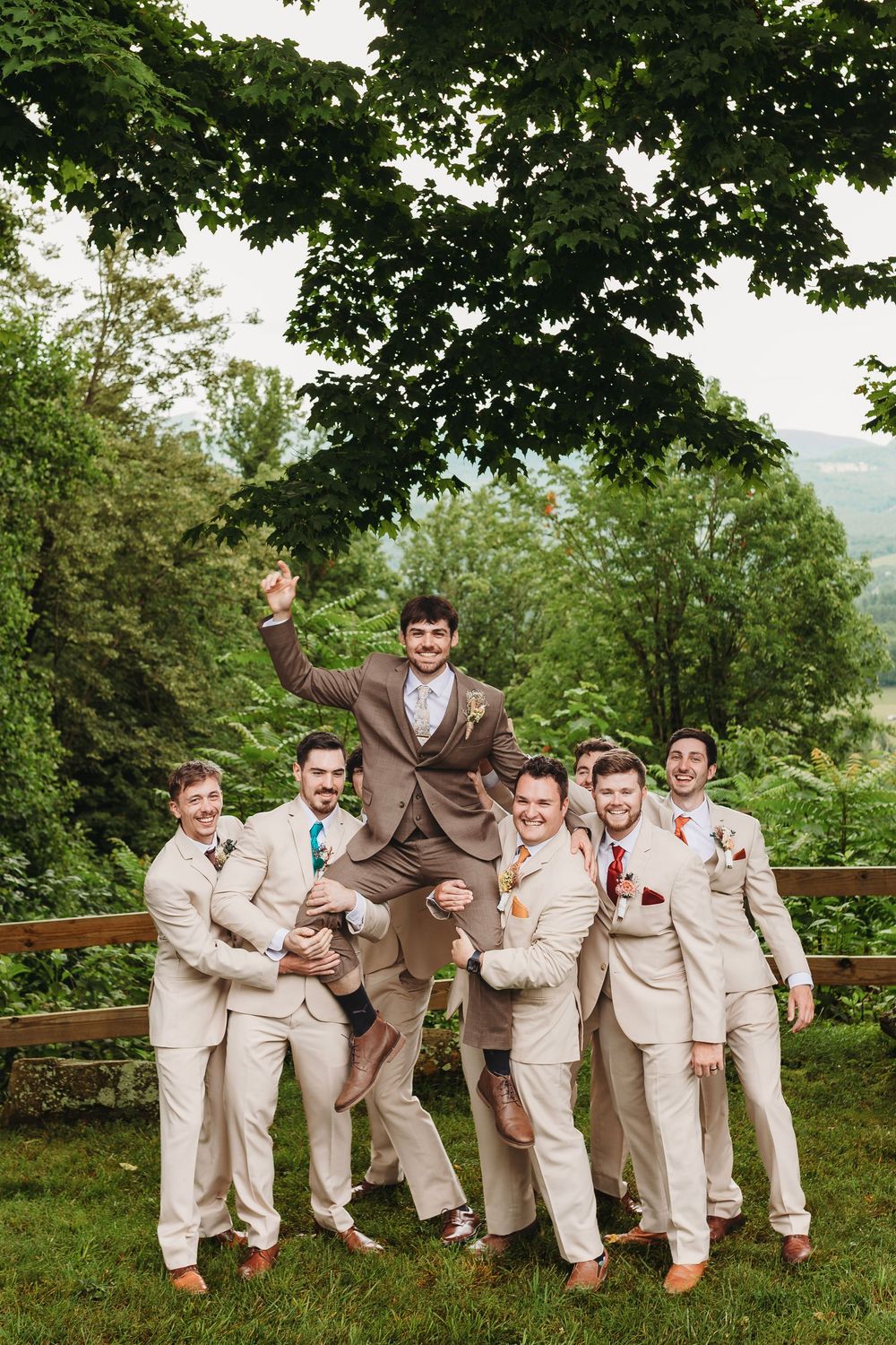 Groom lifted by groomsmen in tan suits outdoors at lush green wedding venue.