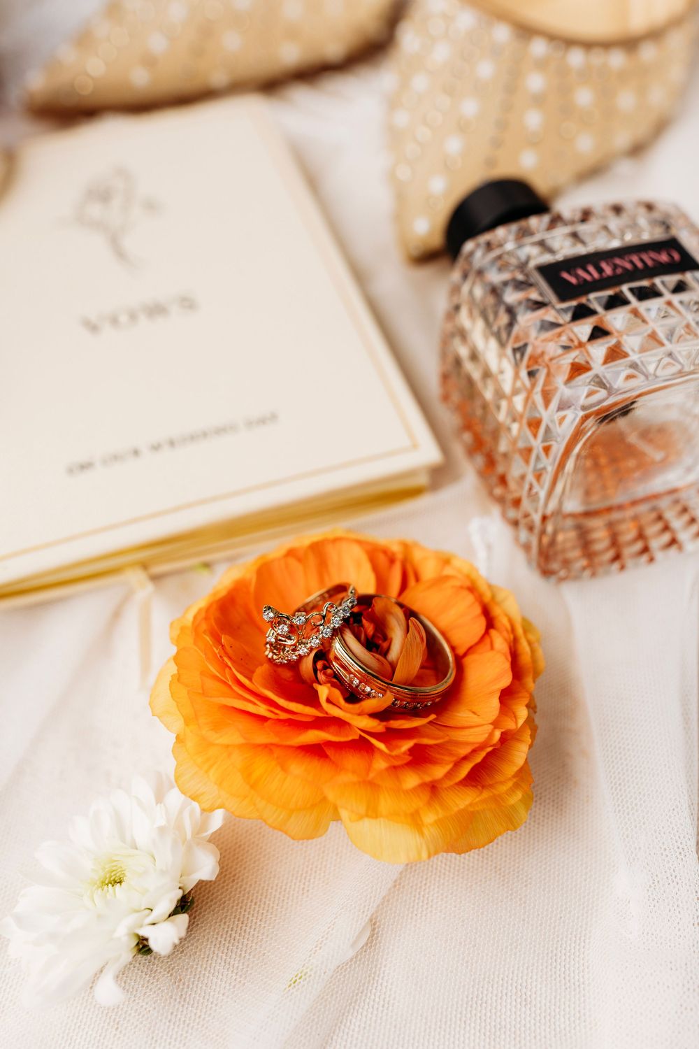 Wedding rings on orange flower with Valentino perfume and vows book on white fabric.