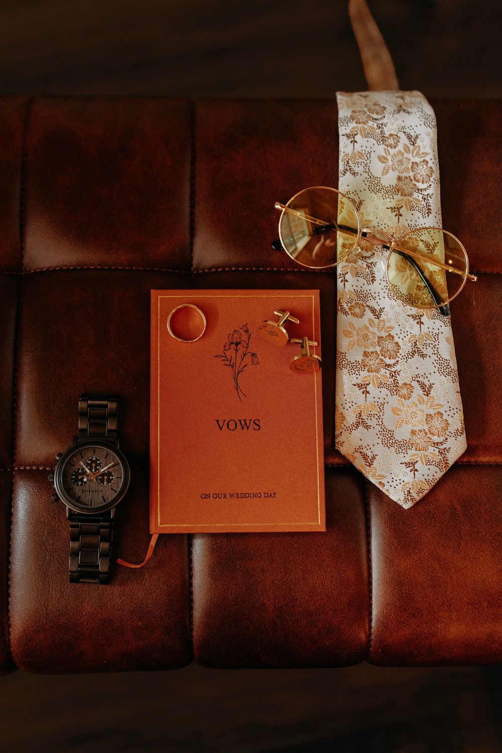 Groom wedding flat lay with vows book, wooden watch, gold cufflinks, ring, floral tie, and round glasses on leather.