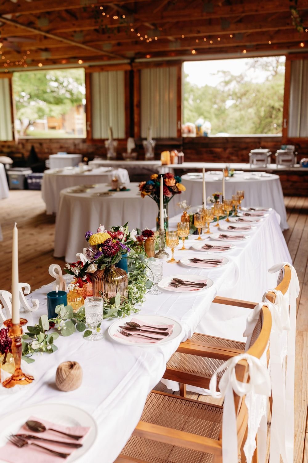 Elegant wedding reception table with white linen, colorful floral centerpieces, candles, and place settings in a rustic barn venue.