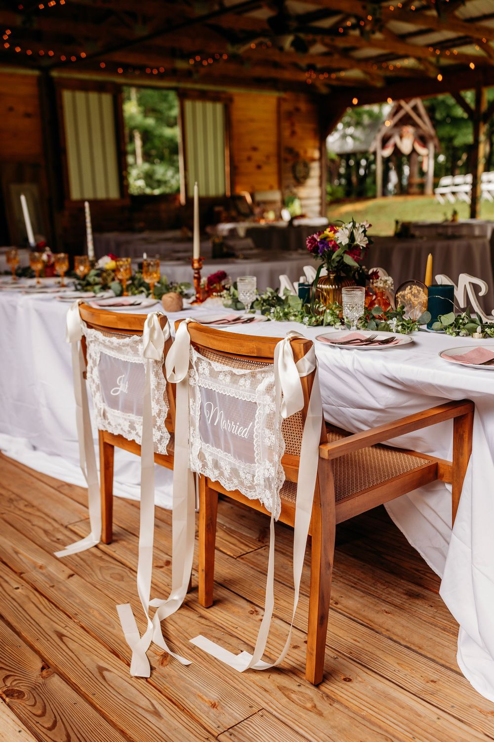 Elegant wedding reception sweetheart table with Just Married chairs, floral centerpieces, and candles in rustic venue.