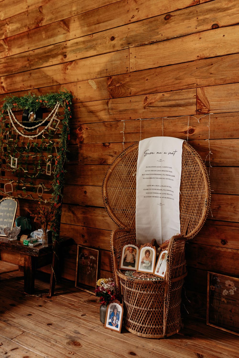 Rustic wooden wall wedding decor with wicker peacock chair, scroll sign, framed photos, and greenery display.