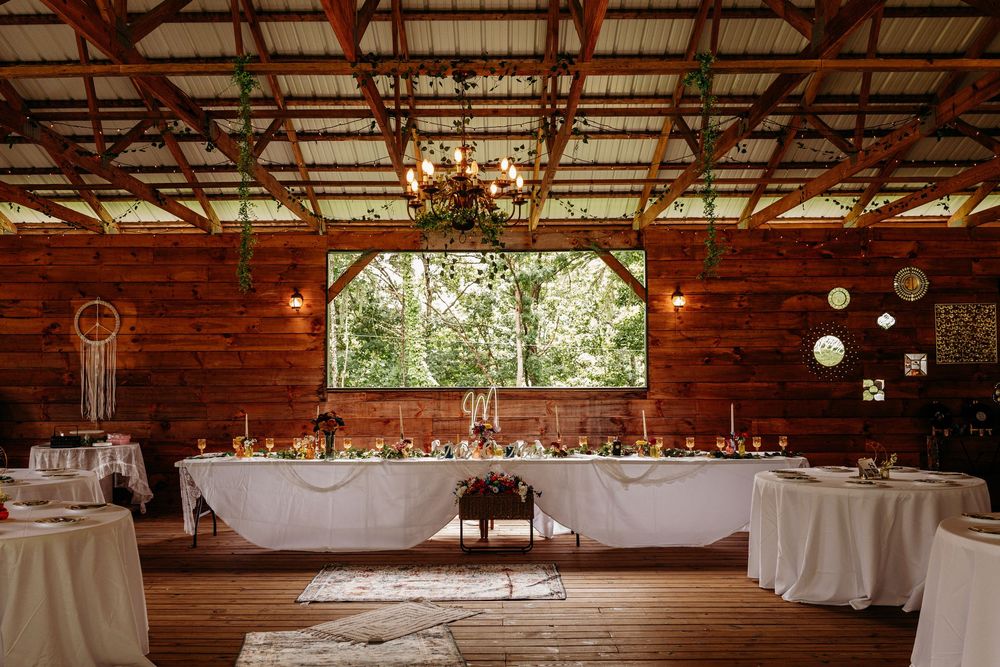 Rustic barn wedding reception with wooden walls, white draped head table, floral centerpieces, and greenery chandelier.