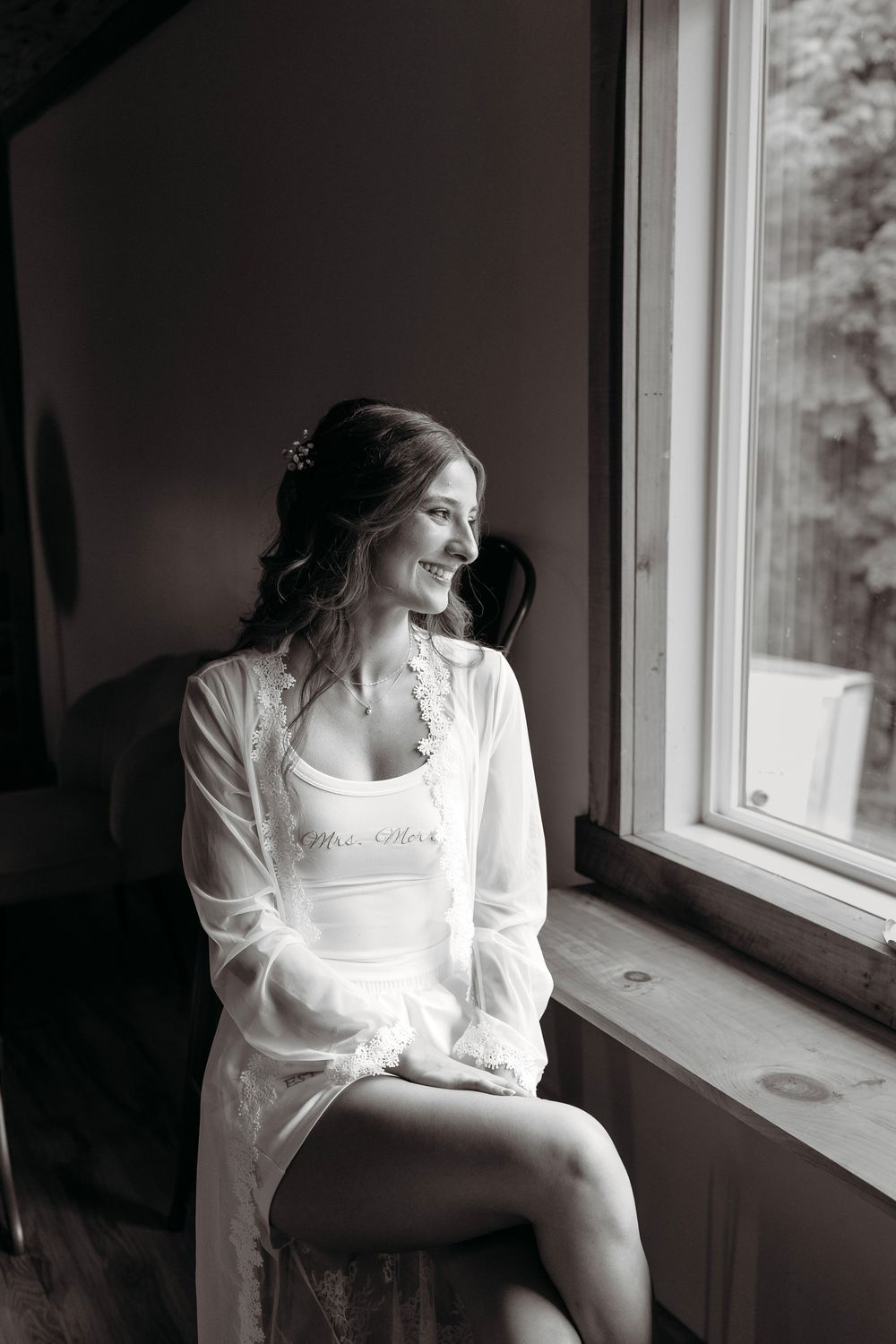 Black and white photo of a smiling bride in a white robe sitting by a window on her wedding day.