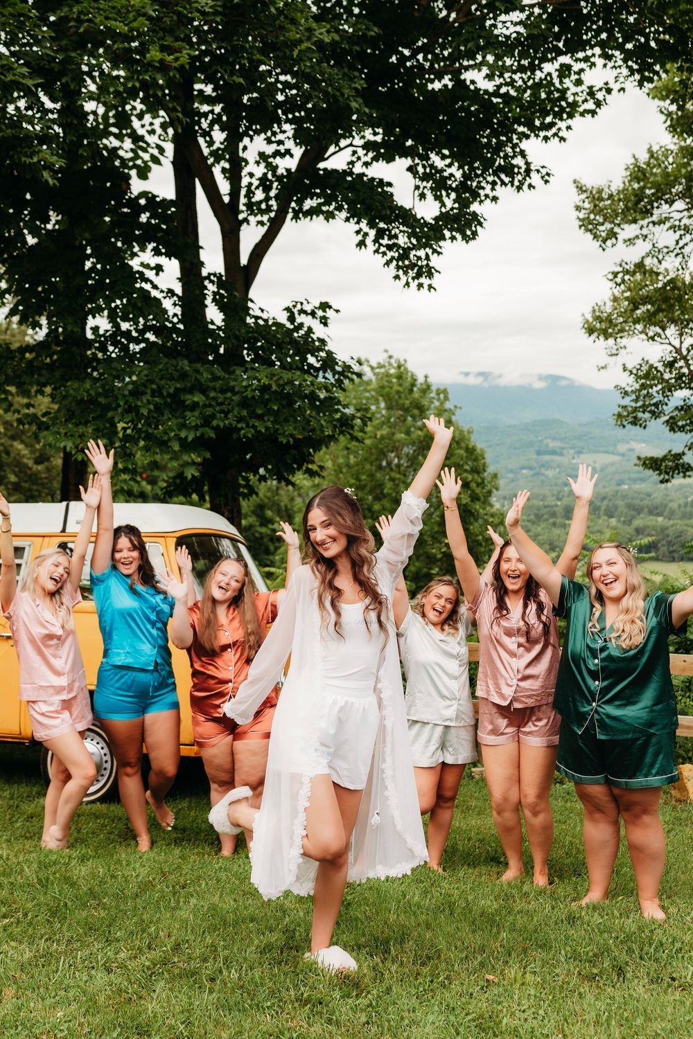 Bride in white robe celebrates with bridesmaids in colorful satin pajamas outdoors near a yellow van with mountain views.