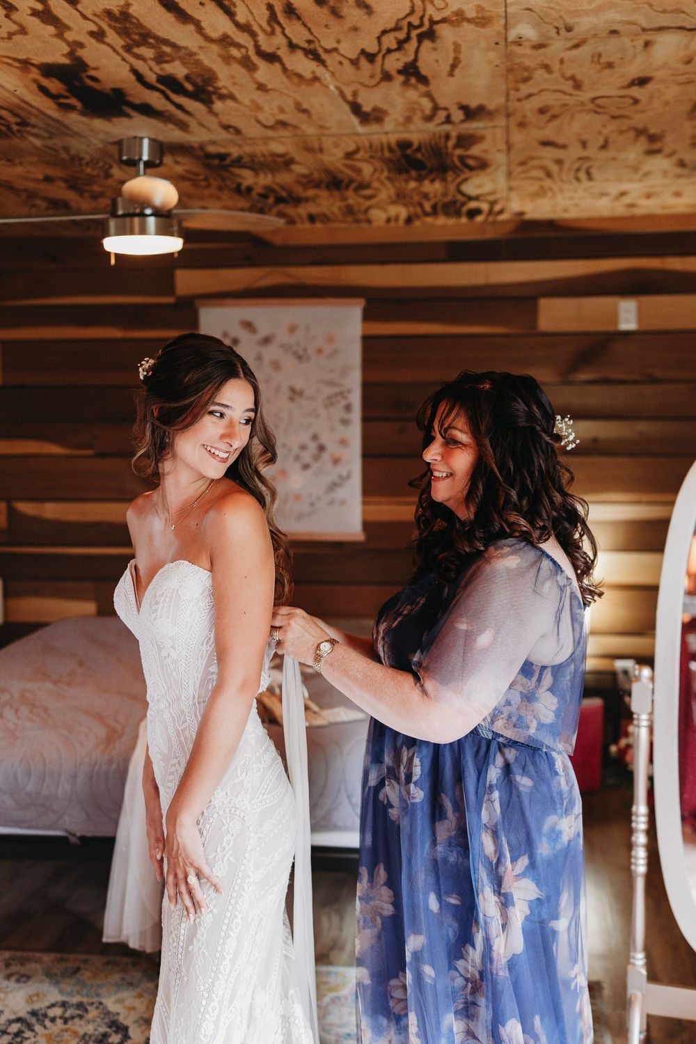 Bride in white lace gown being helped by woman in blue floral dress inside a rustic log cabin bedroom.