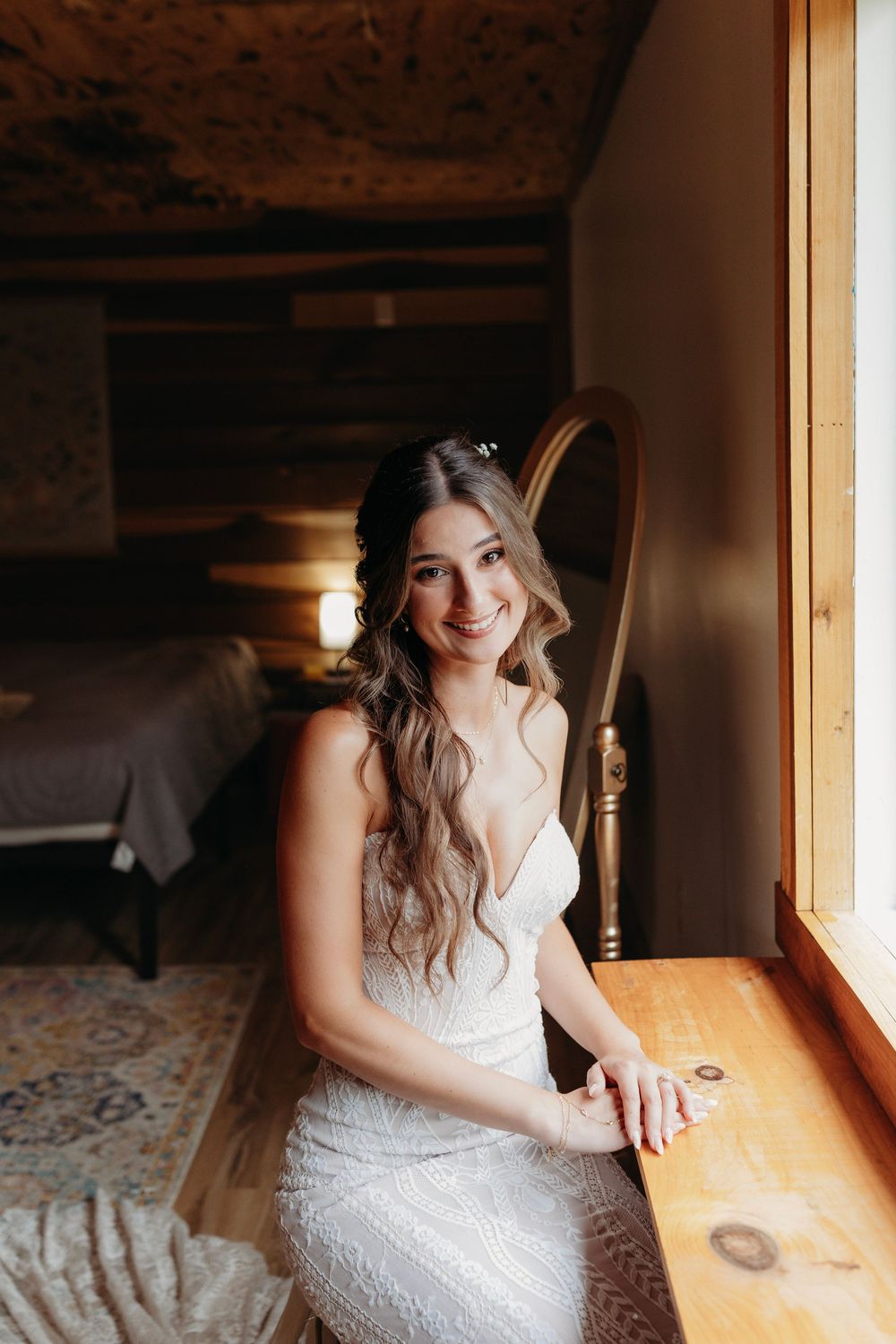 Smiling bride in white lace gown sitting by window in rustic log cabin bedroom with warm lighting.