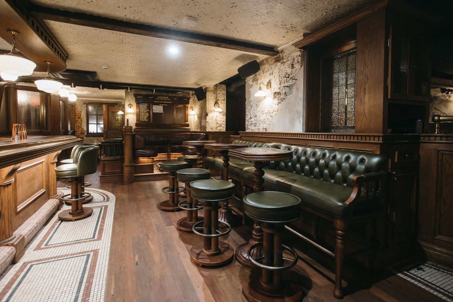 Cozy traditional pub interior with dark wood bar stools, green leather booth seating, and rustic stone walls.