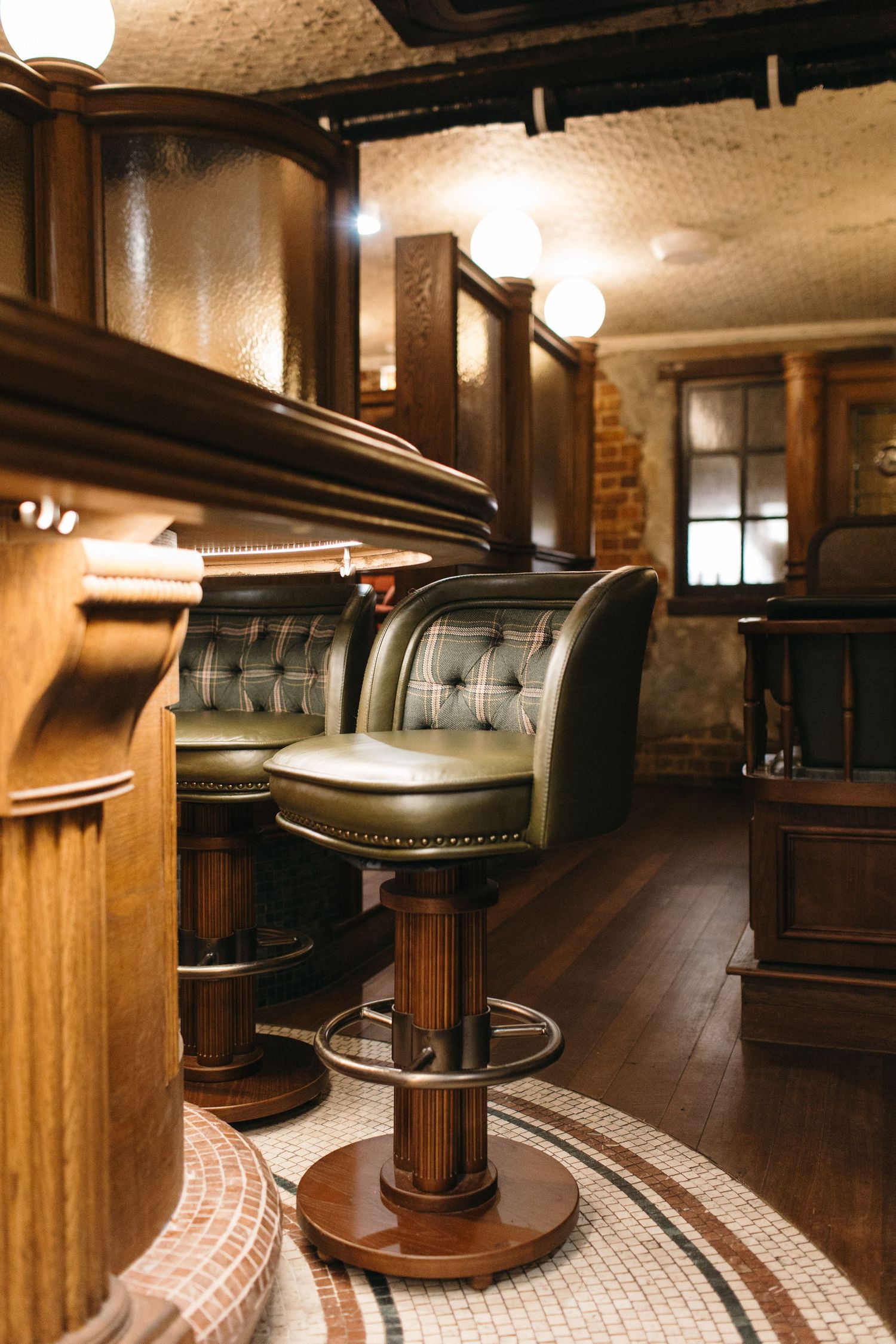 Elegant bar interior with tufted green leather bar stools, dark wood paneling, and warm globe lighting.