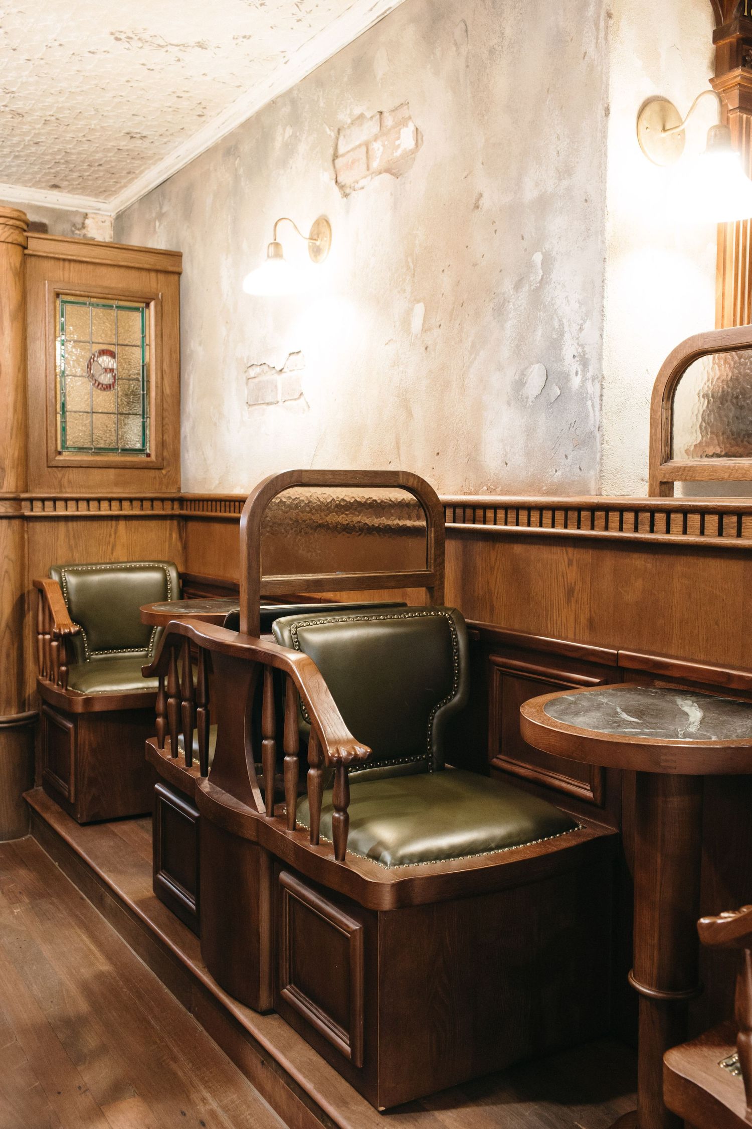 Vintage pub interior with green leather chairs, dark wood paneling, stained glass window, and antique wall sconces.