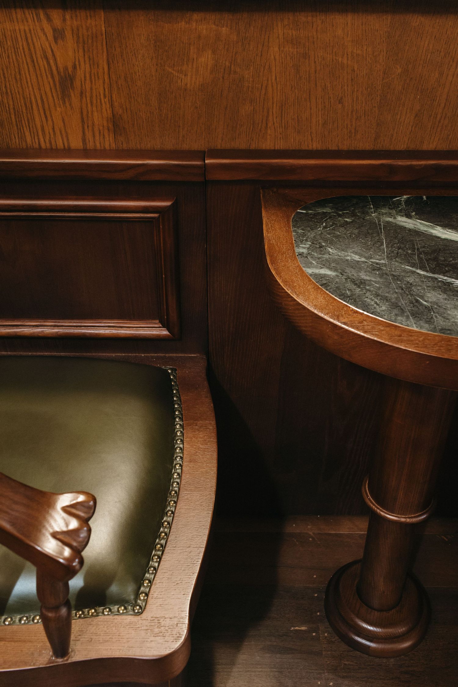 Elegant wooden furniture set featuring a green leather chair and marble-top pedestal side table against wood paneling.