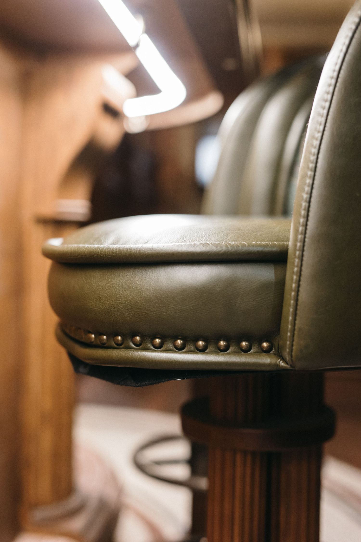 Close-up of a green leather bar stool with brass nail head trim and ribbed wooden base in a warmly lit interior.