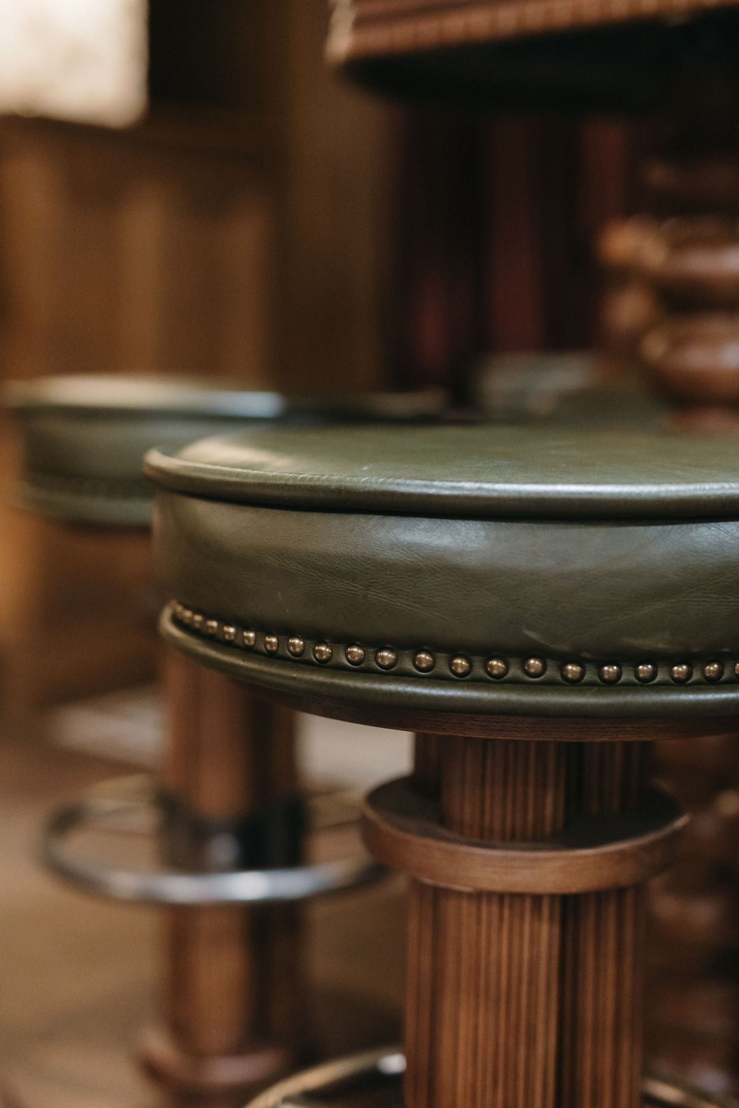 Green leather bar stools with brass nail head trim on wooden pedestals in a classic bar setting.