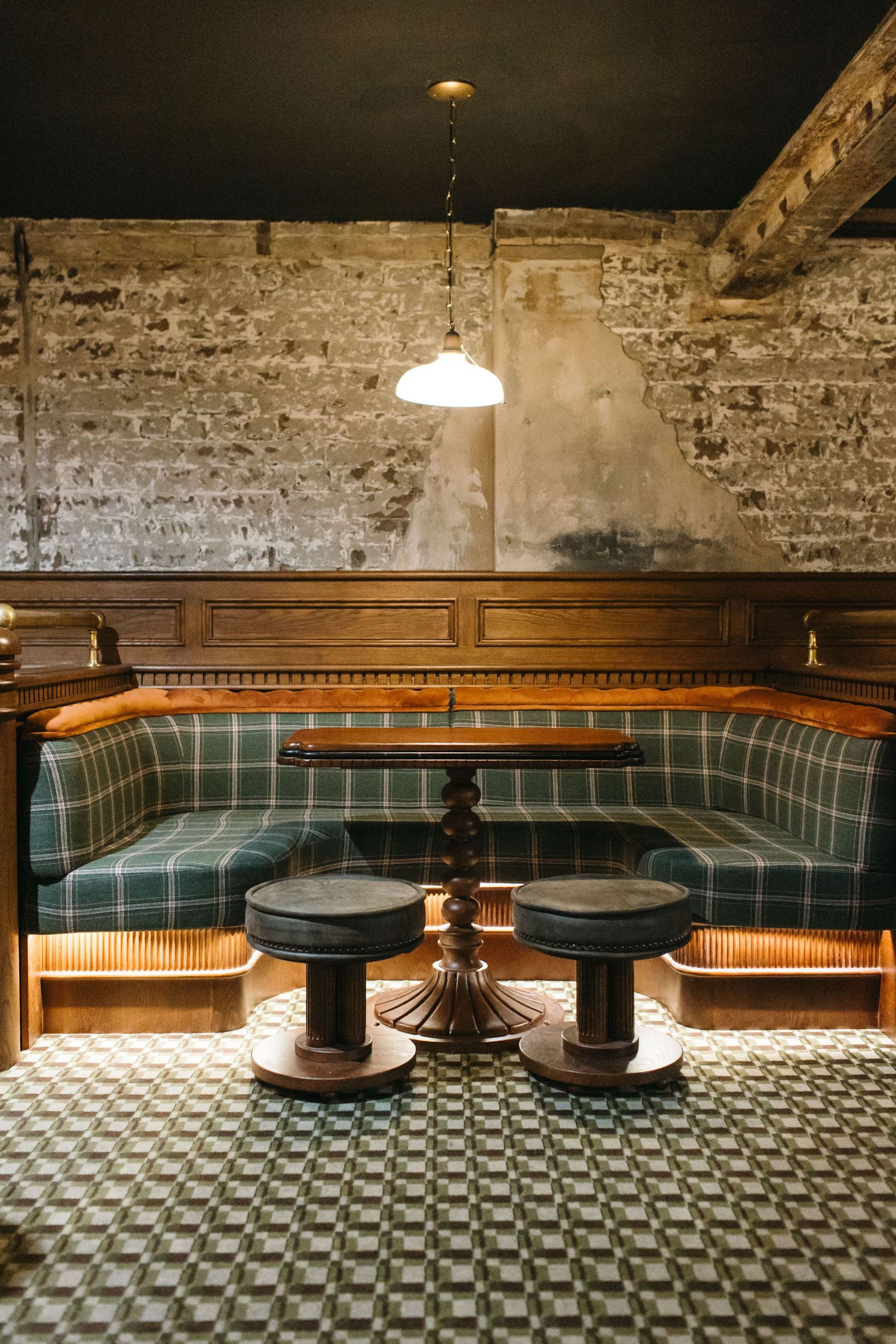 Cozy restaurant booth with green plaid upholstery, wooden paneling, two stools, and a pendant light against a brick wall.