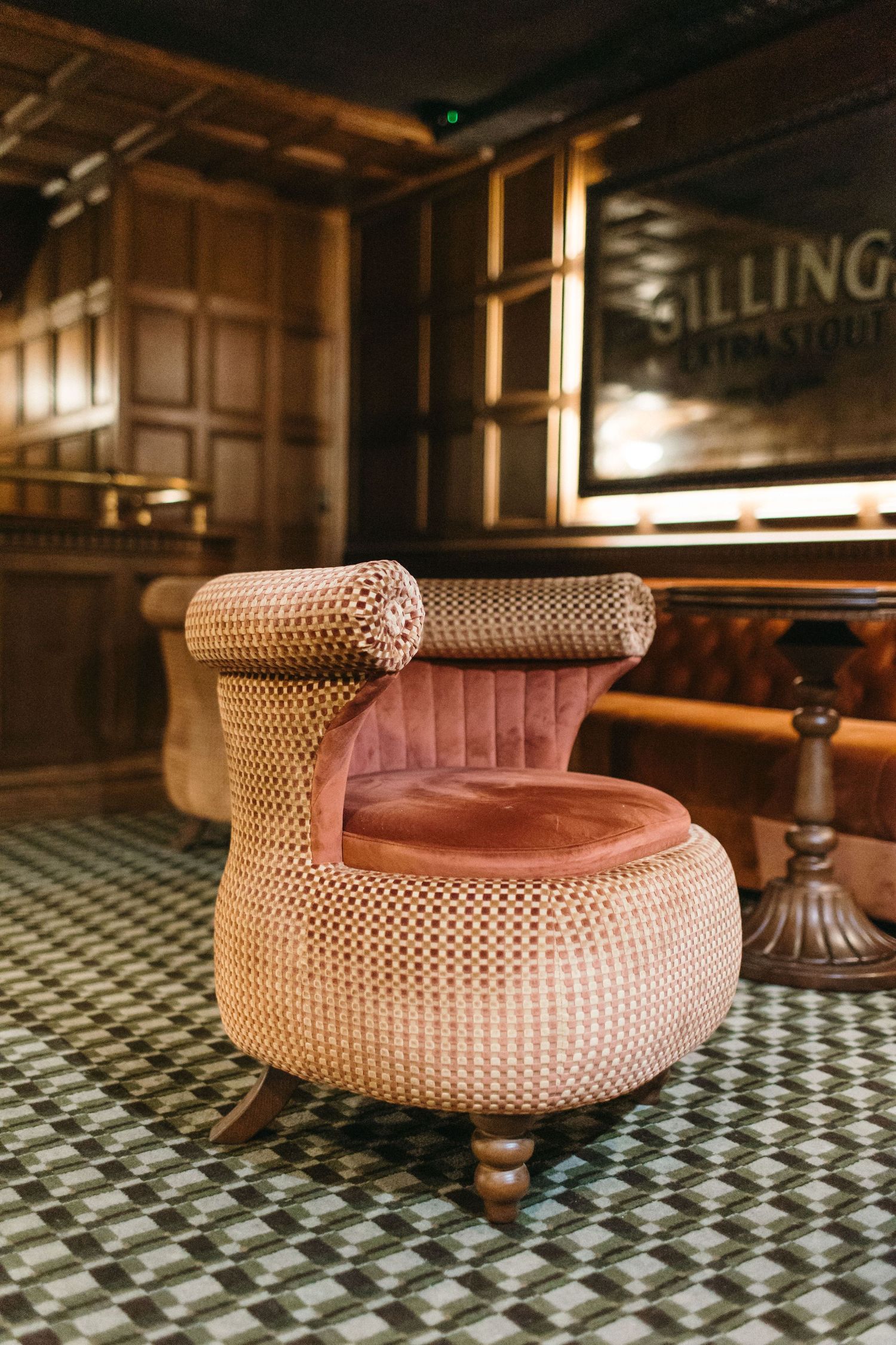 A vintage rattan armchair with pink velvet cushions on a geometric tile floor in an elegant wood-paneled bar.