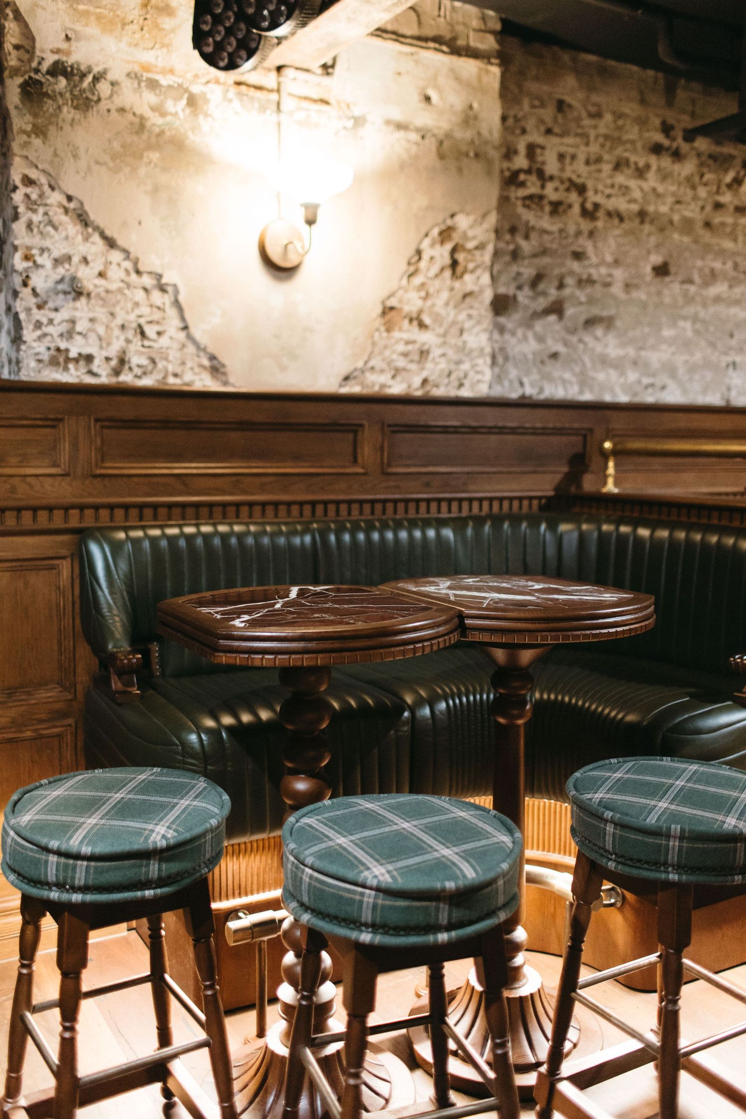 Cozy pub corner with green leather booth, plaid bar stools, and wooden tables against a rustic stone wall.