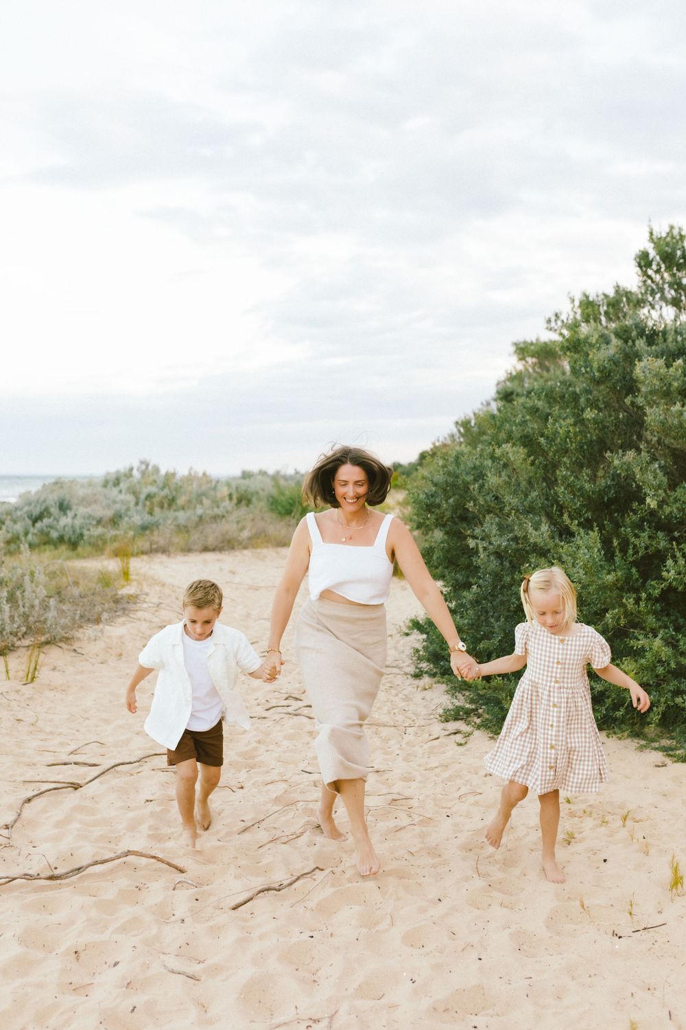 A mother walks barefoot on sandy beach path holding hands with two young children on a cloudy day.