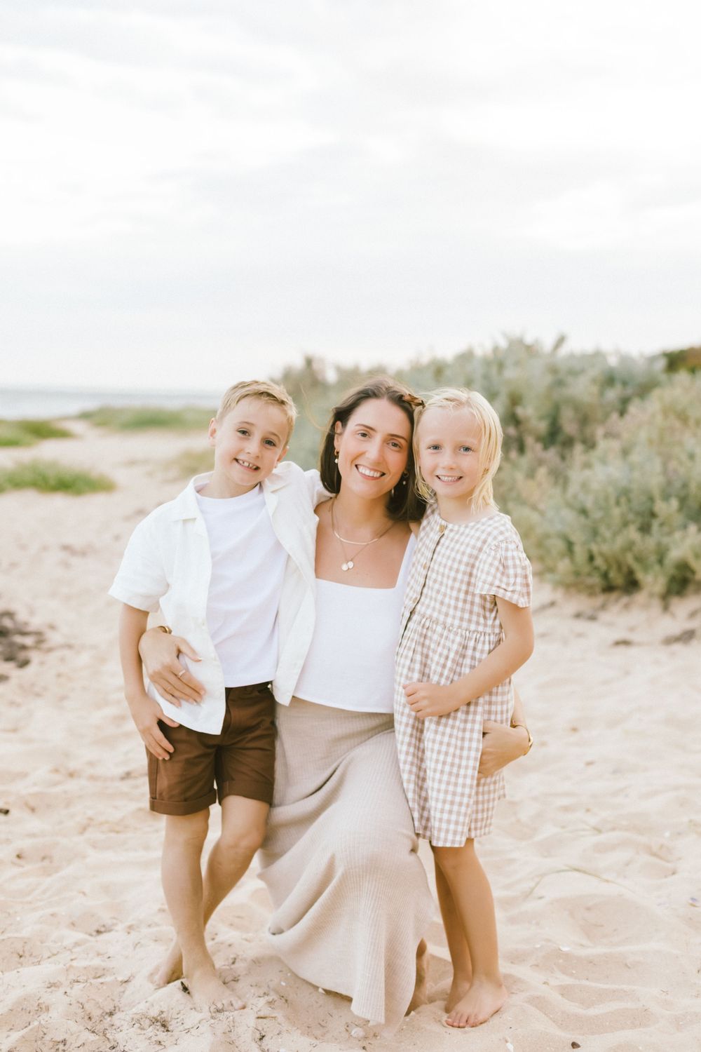 A smiling woman poses with two young children on a sandy beach, wearing casual summer outfits on a cloudy day.
