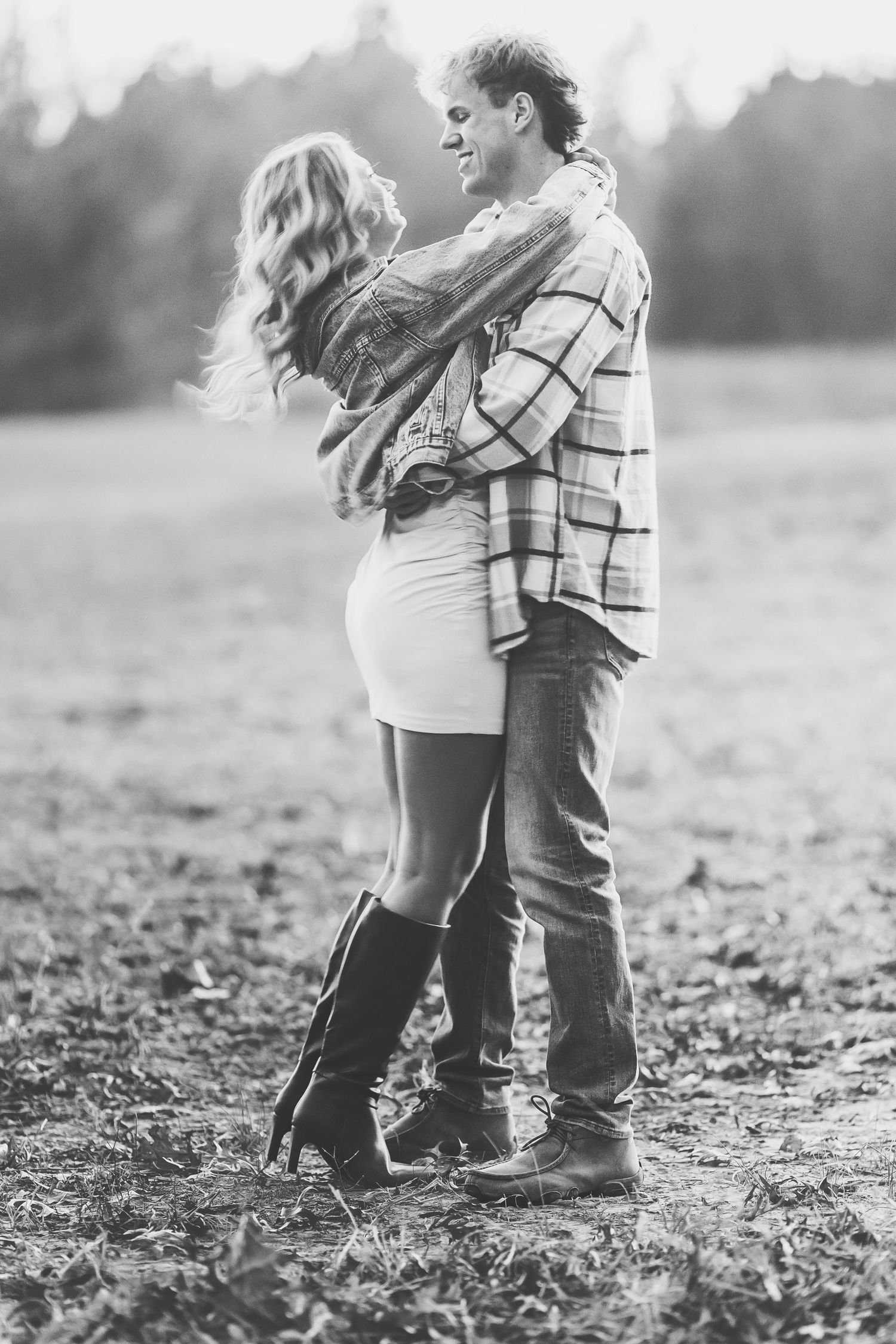 Black and white photo of a romantic couple embracing in an autumn field, woman in boots and short dress.