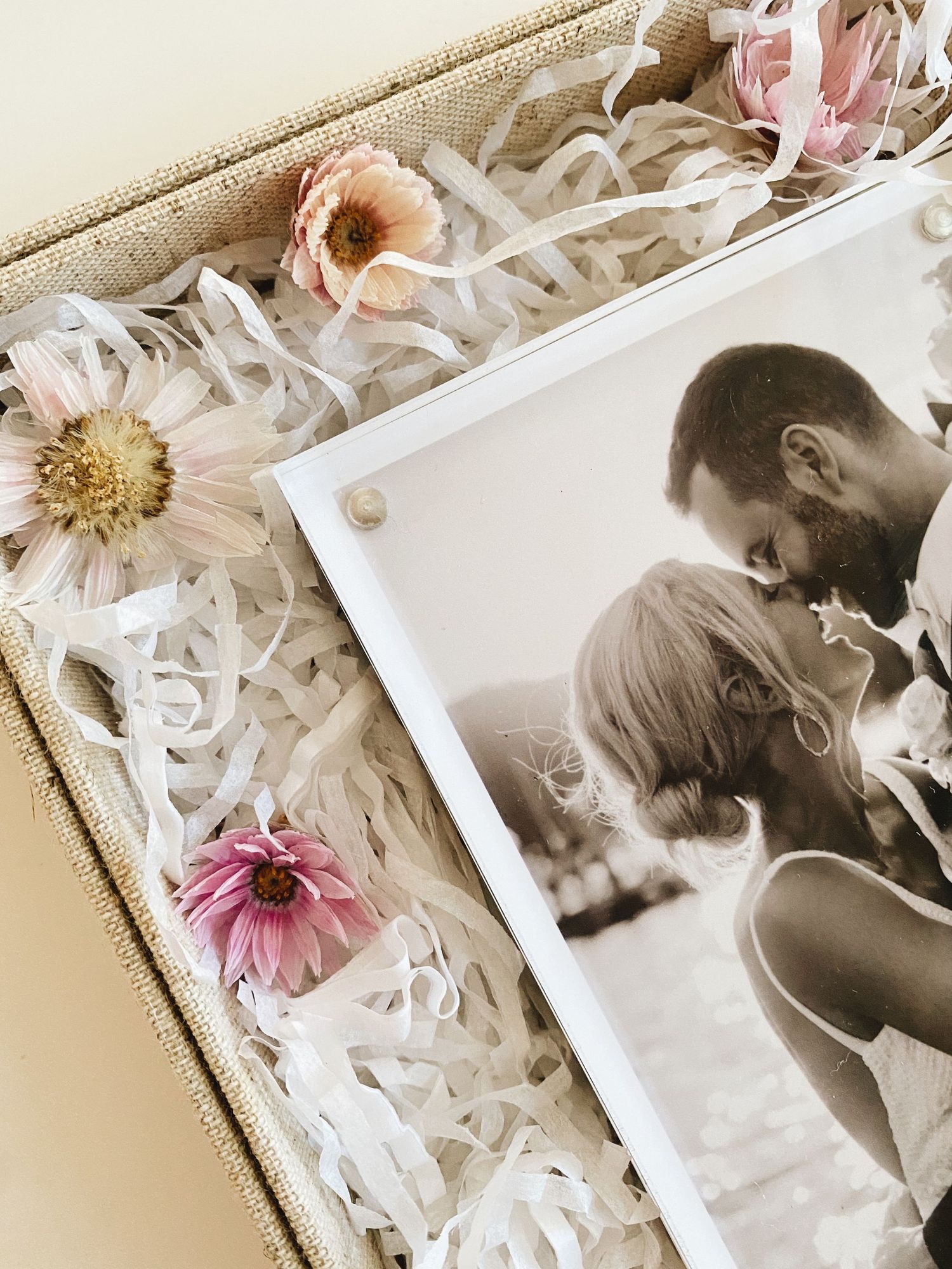 A romantic black and white wedding photo in an acrylic frame, nestled in a gift box with shredded paper and pink daisy flowers.