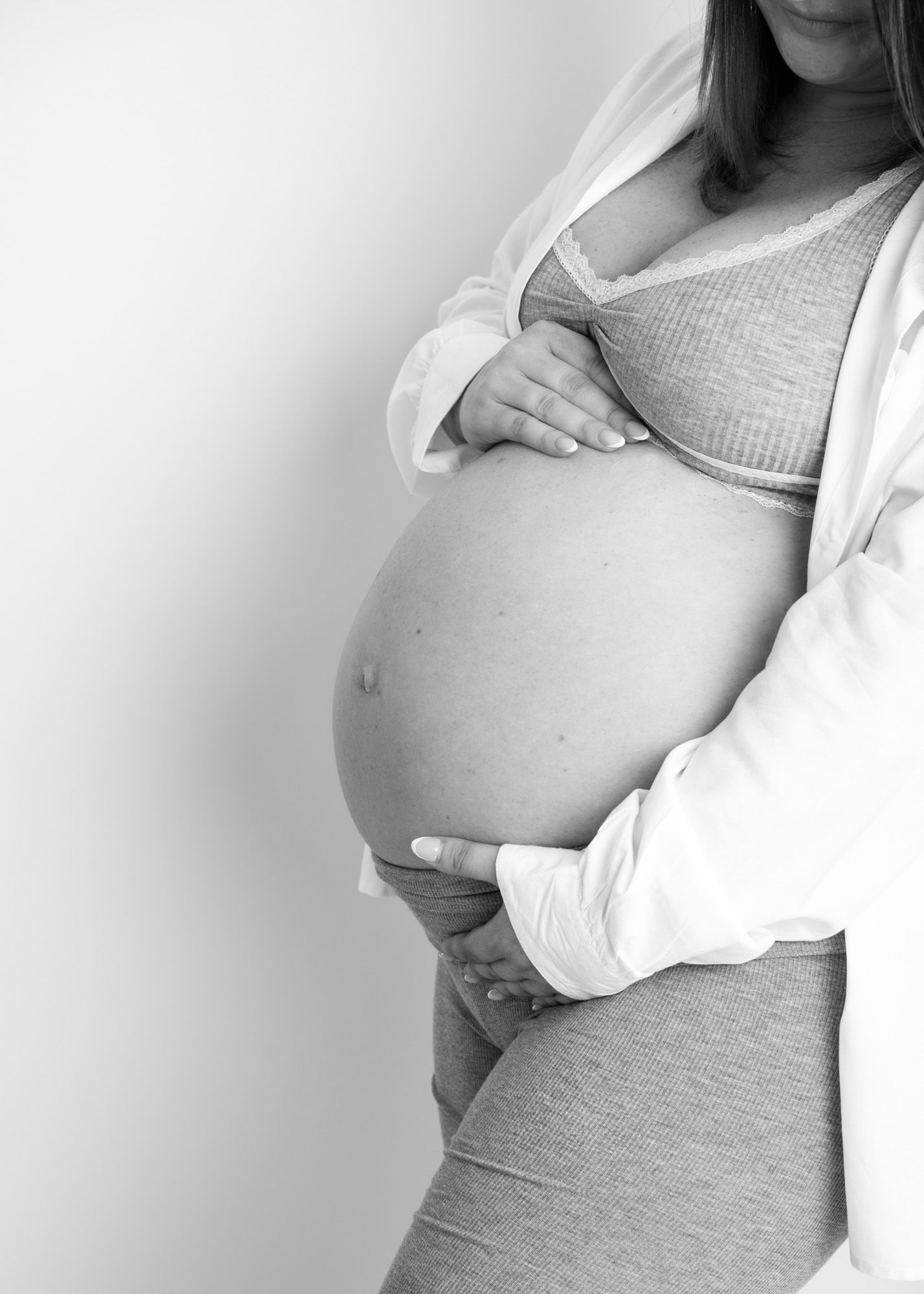 Black and white photo of a pregnant woman cradling her bare baby bump, wearing a cozy crop top and open cardigan.