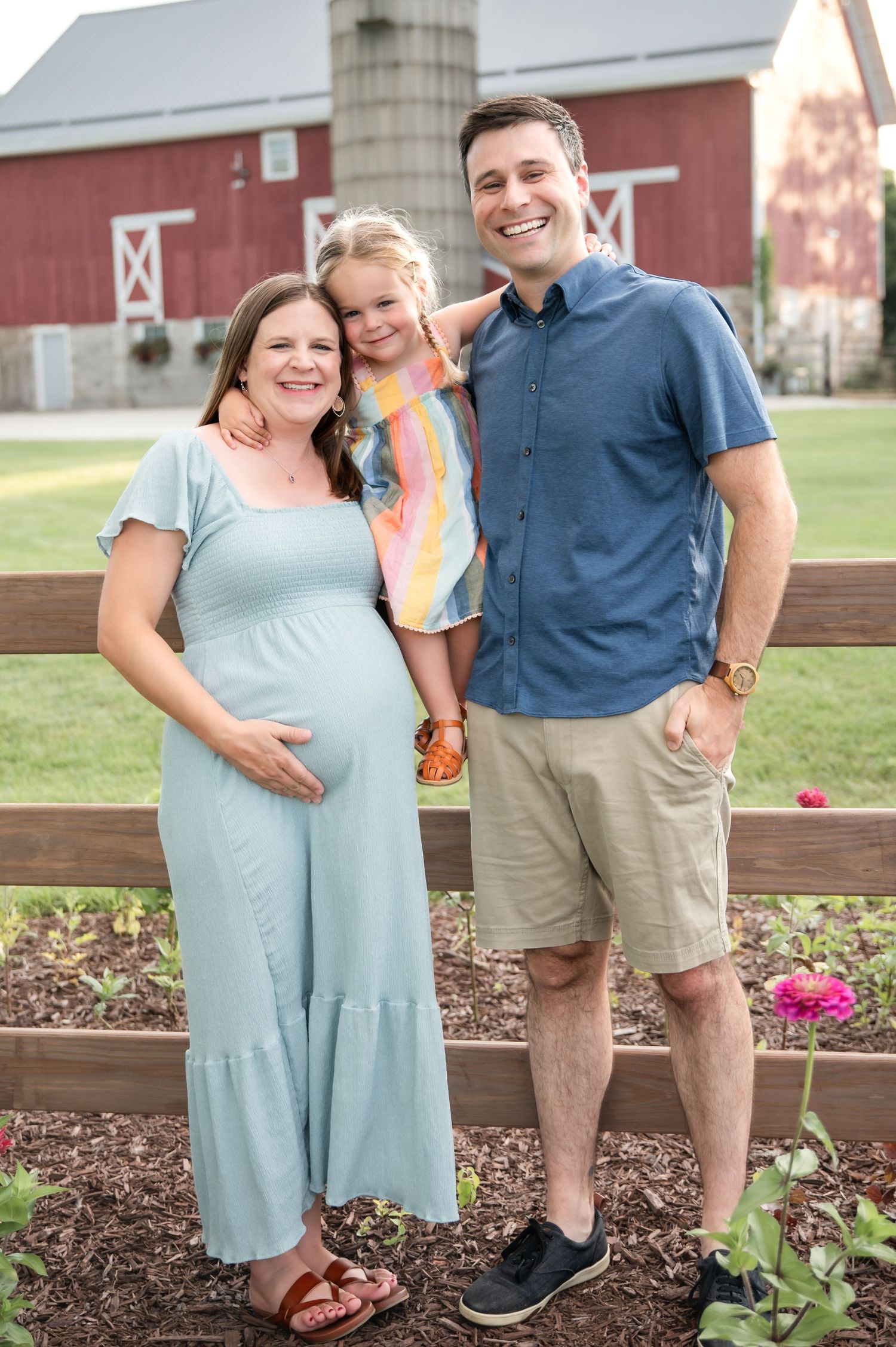 Smiling pregnant woman in mint dress with husband and young daughter posing outdoors near a red barn and flower garden.