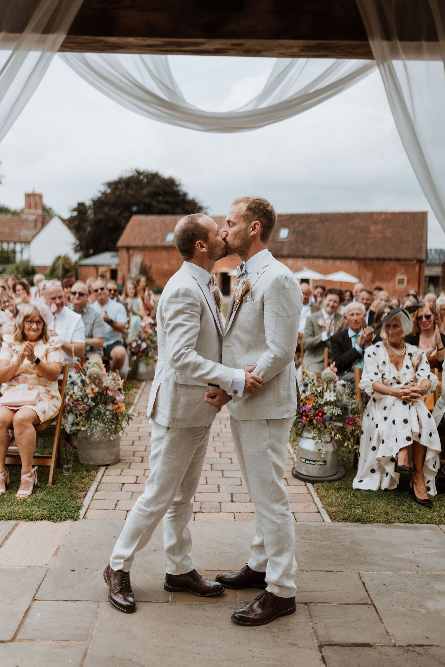 Two grooms in matching light suits share their first kiss at an outdoor wedding ceremony surrounded by guests at Moat Hall Barns, Shrewsbury